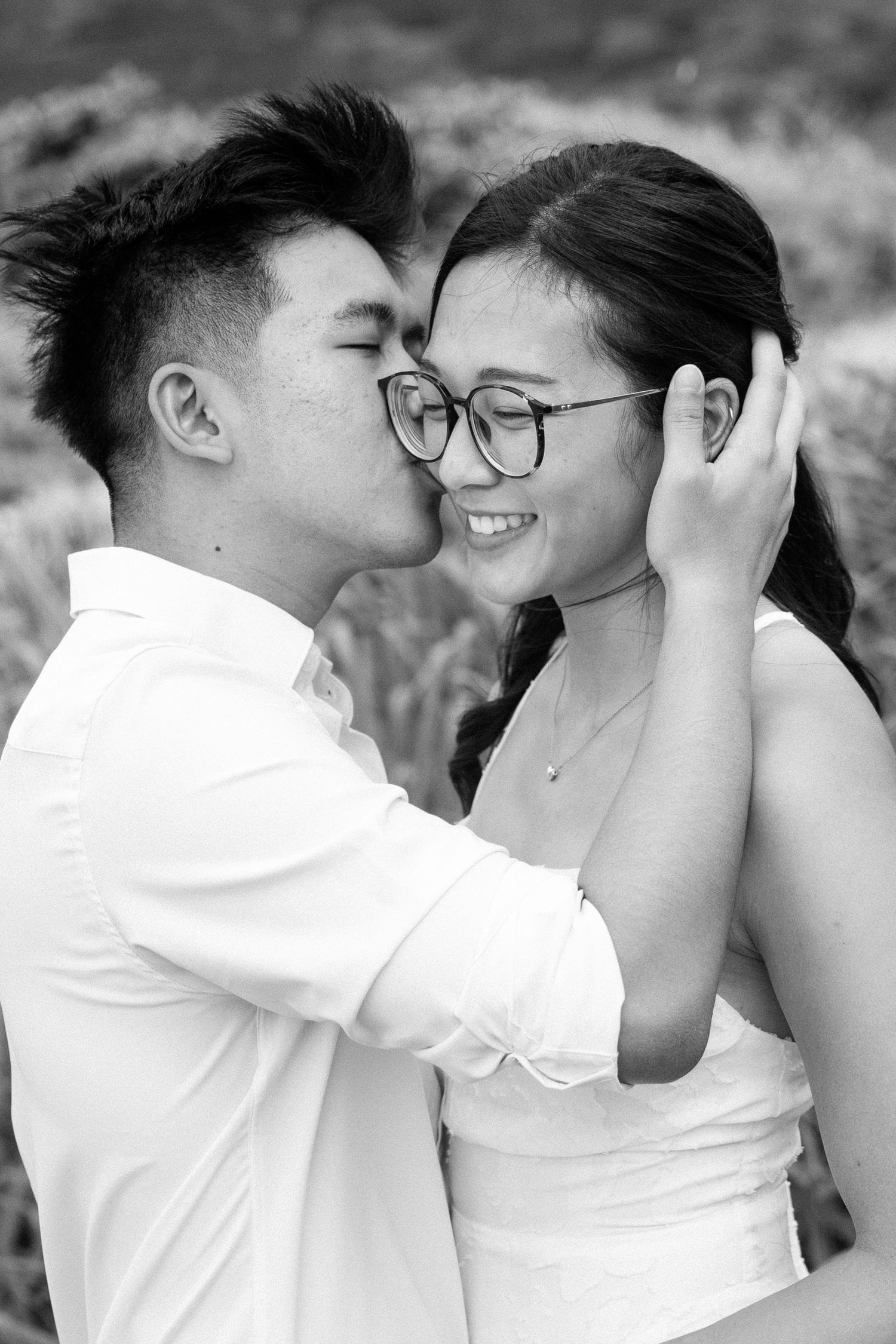 Couple sharing a tender kiss in black and white engagement photo, woman smiling with glasses as man kisses her cheek.