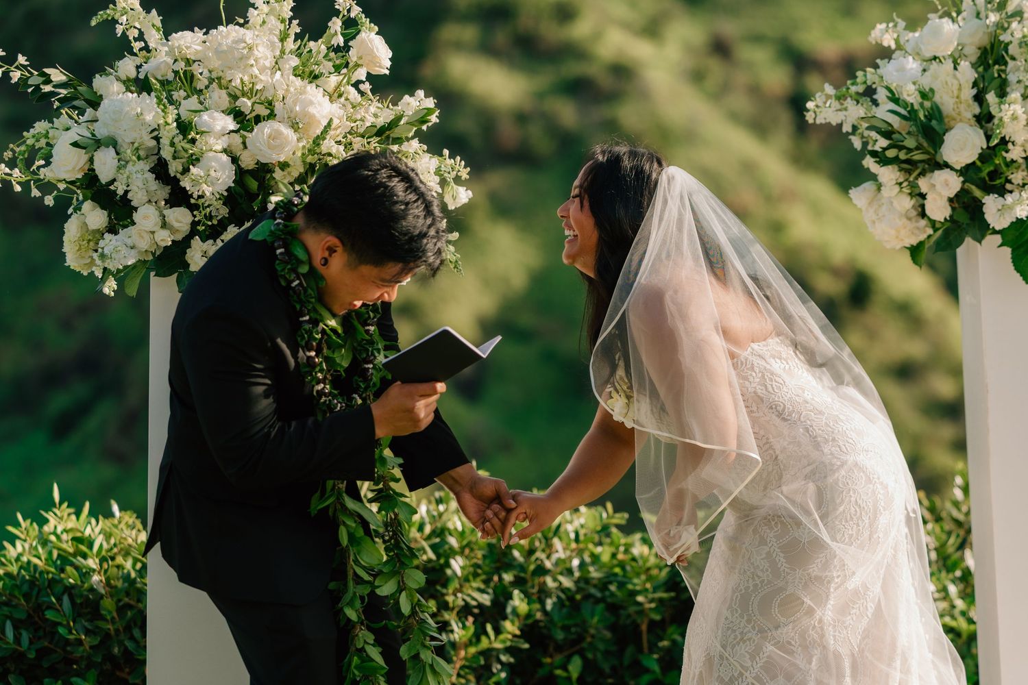 Couple exchanging wedding vows outdoors under white floral arch with lush green hills backdrop.