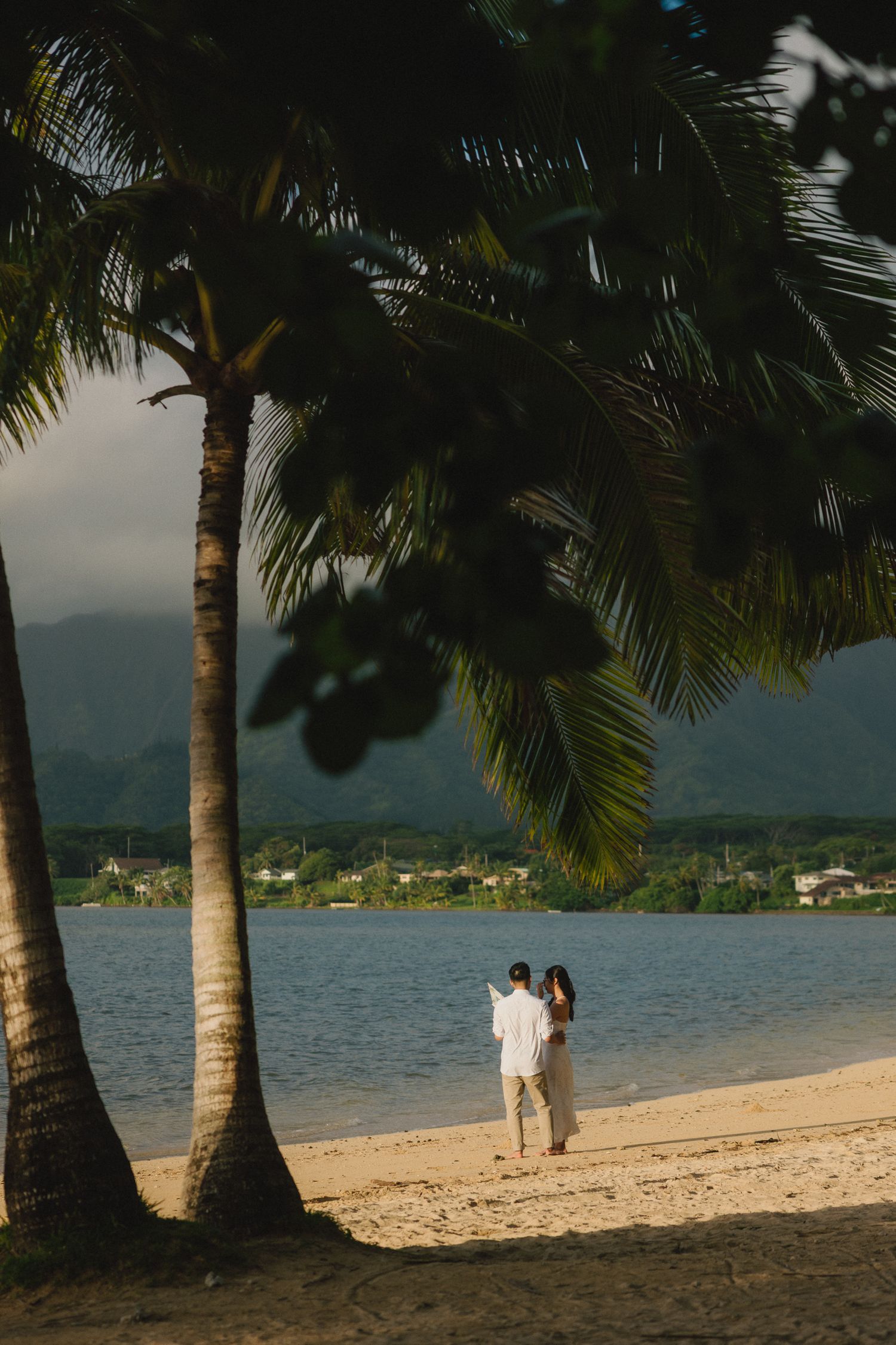 Couple walking hand in hand on a tropical beach with palm trees and mountain views in the background.