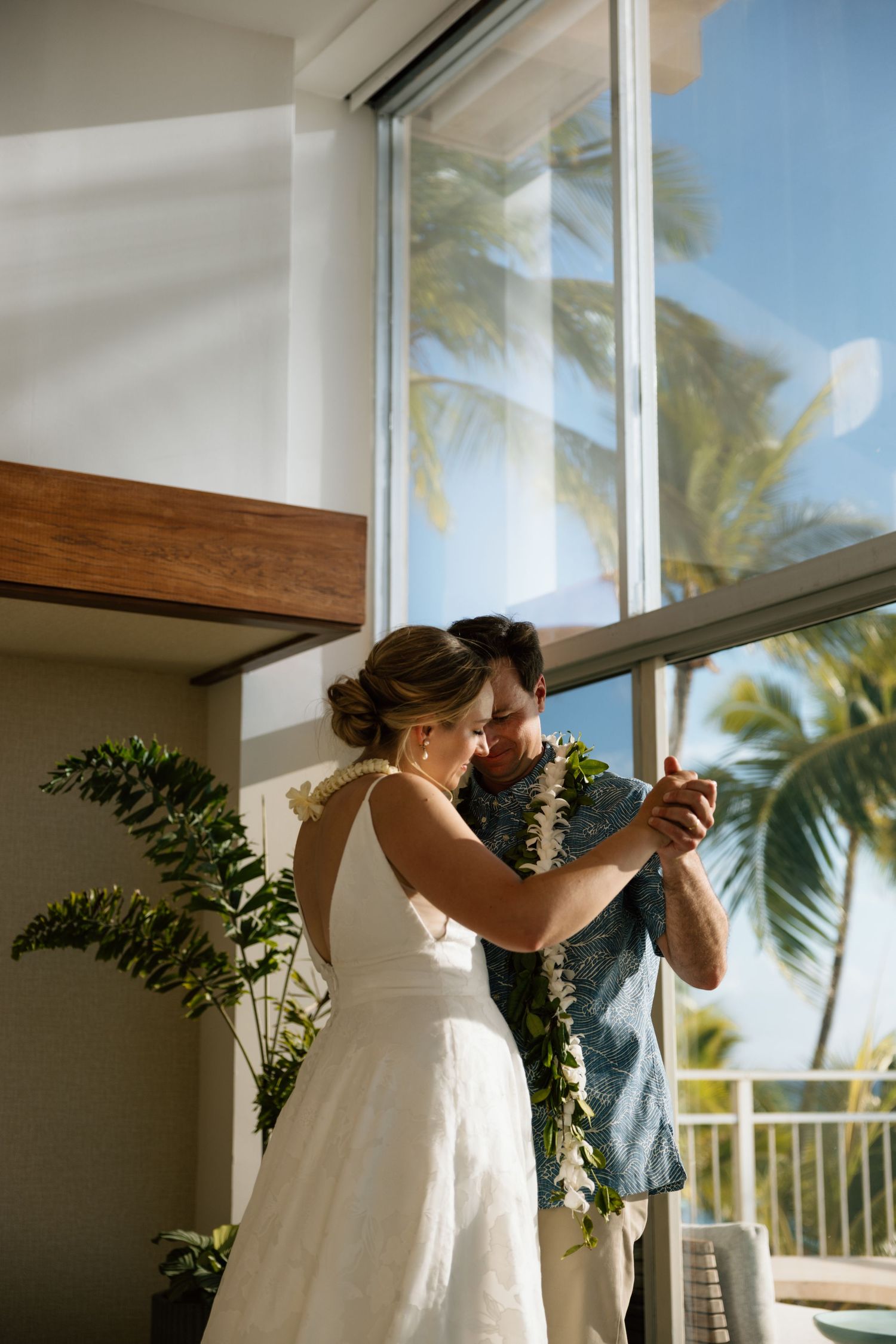 Couple sharing a first dance indoors with tropical palm trees visible through large windows.