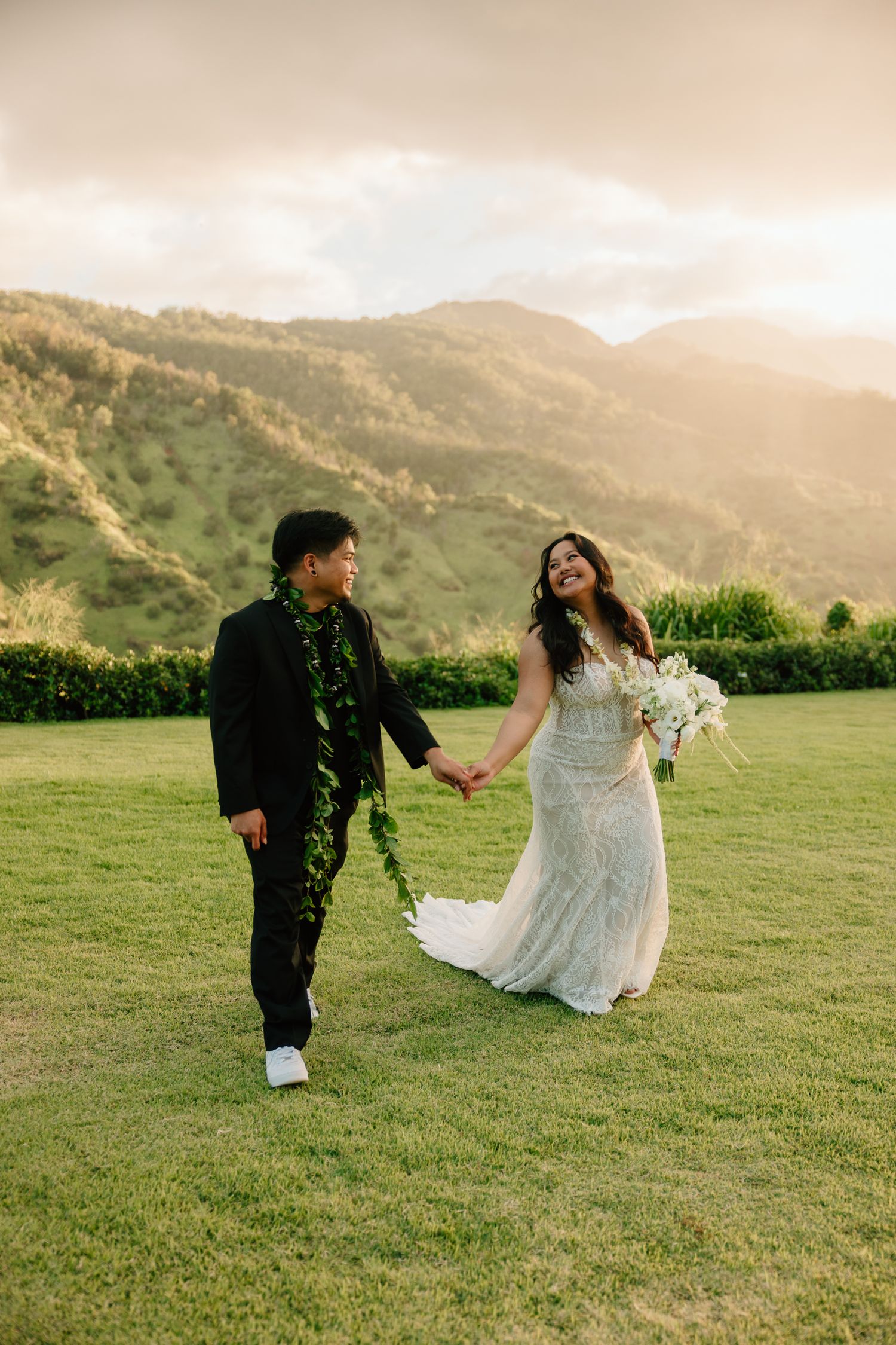 Newlyweds walk hand-in-hand at sunset on green lawn with mountains, groom wearing Hawaiian lei, bride in lace gown.