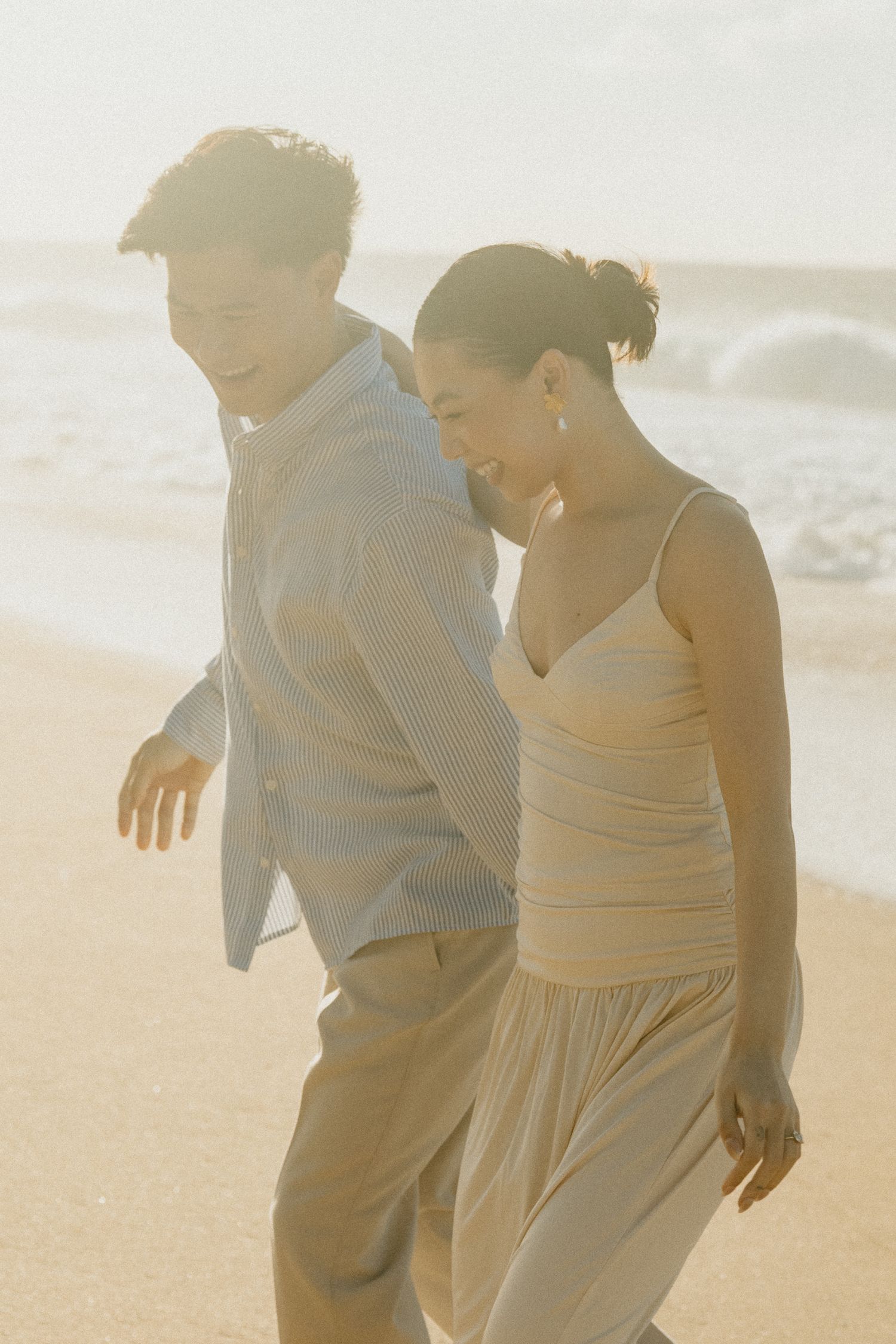 Couple walking together on a sunlit beach with ocean waves in the soft golden background.