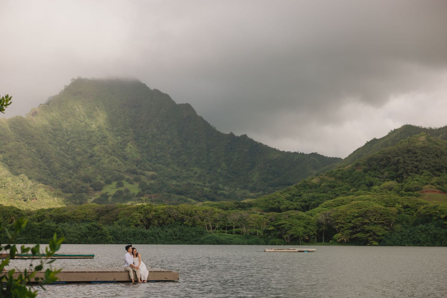 Couple sitting on a dock by a serene lake with lush green mountains and dramatic cloudy skies in Hawaii.