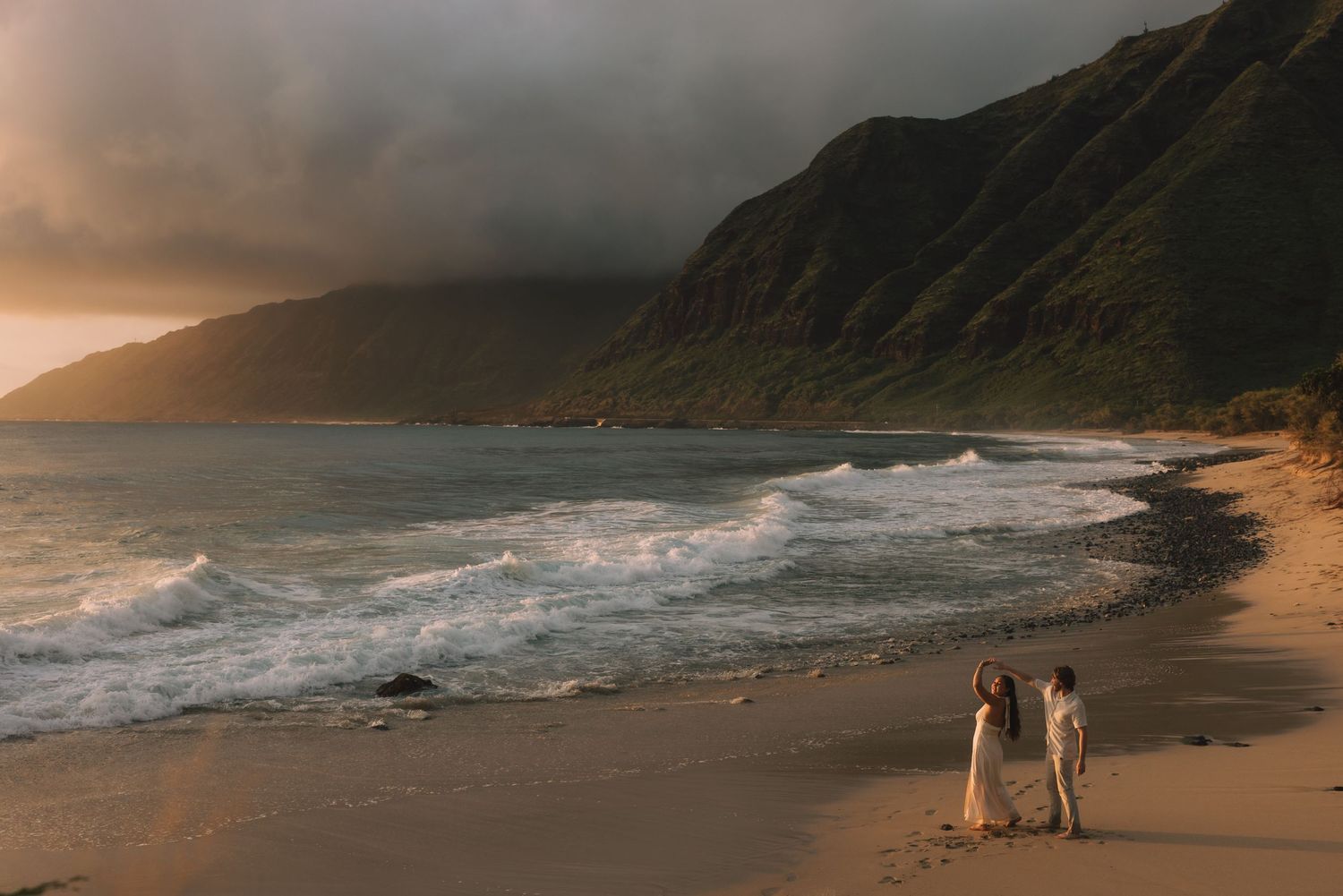 Couple walks hand in hand on a golden sandy beach at sunset with dramatic stormy skies and lush green mountains.