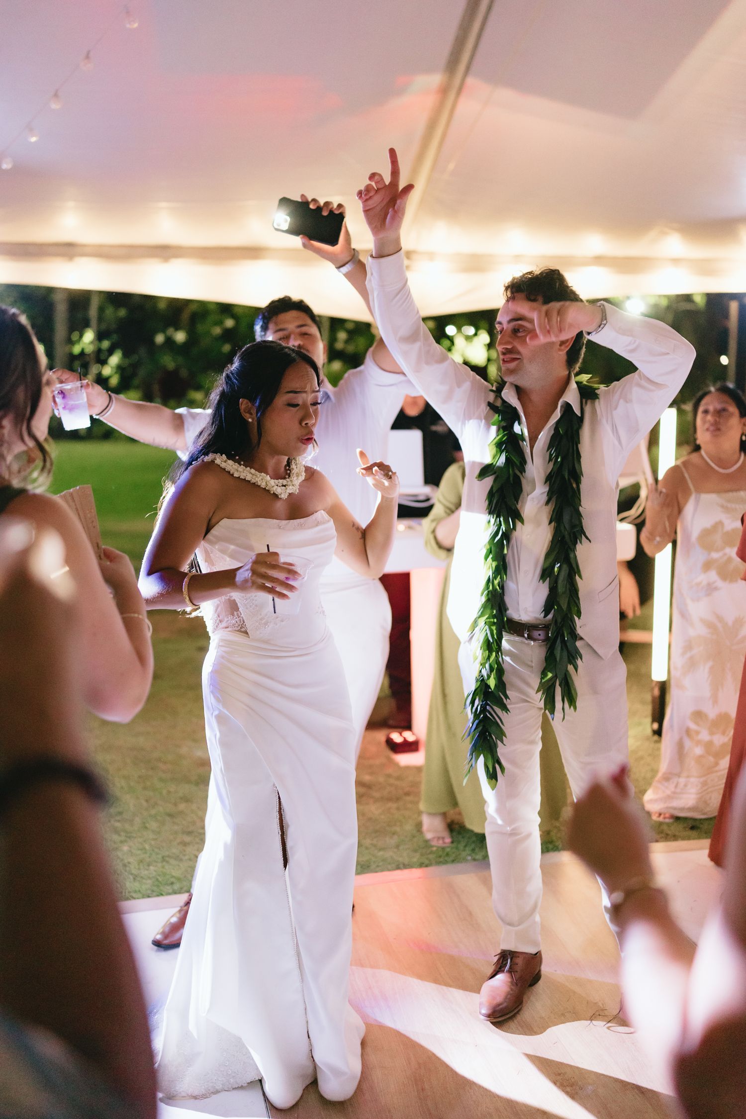 Bride and groom dancing joyfully at outdoor Hawaiian wedding reception under string lights, groom wearing green lei.
