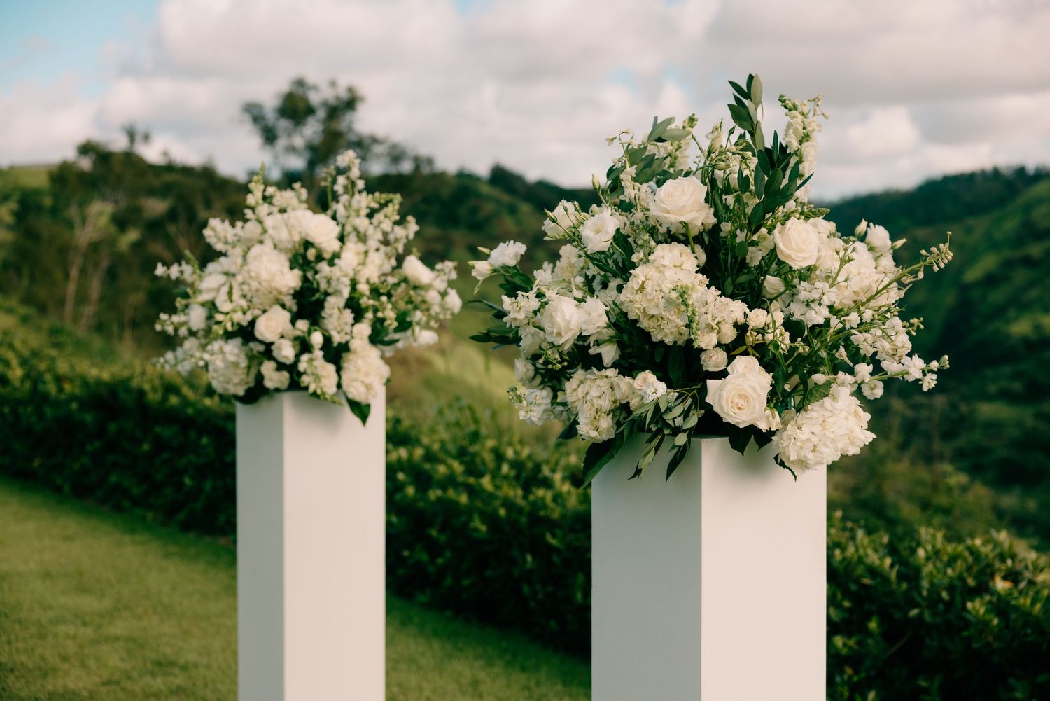 Two white pedestals topped with lush white floral arrangements at an outdoor wedding ceremony with green hills backdrop.