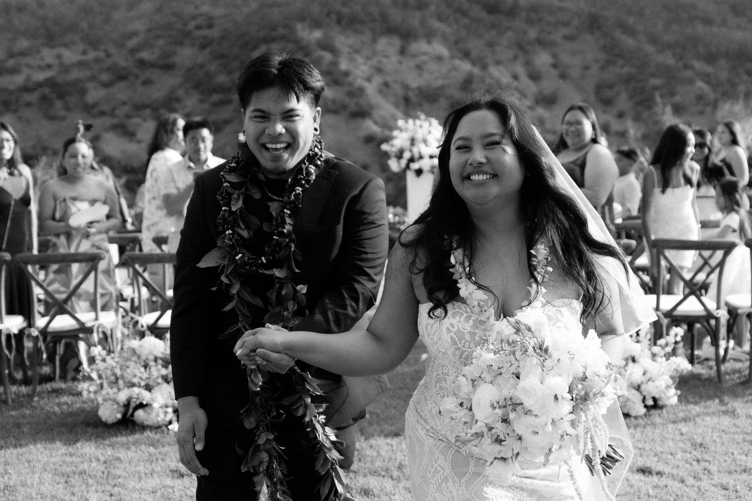 Black and white photo of a smiling couple in wedding attire walking outdoors after their ceremony.