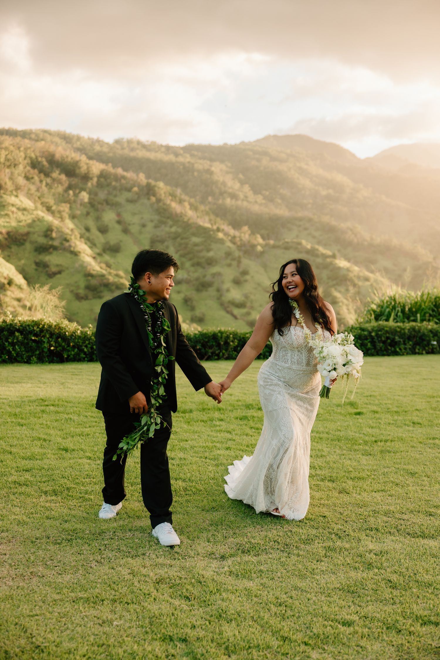 Bride and groom holding hands walking on green lawn with lush Hawaiian mountains at golden hour.