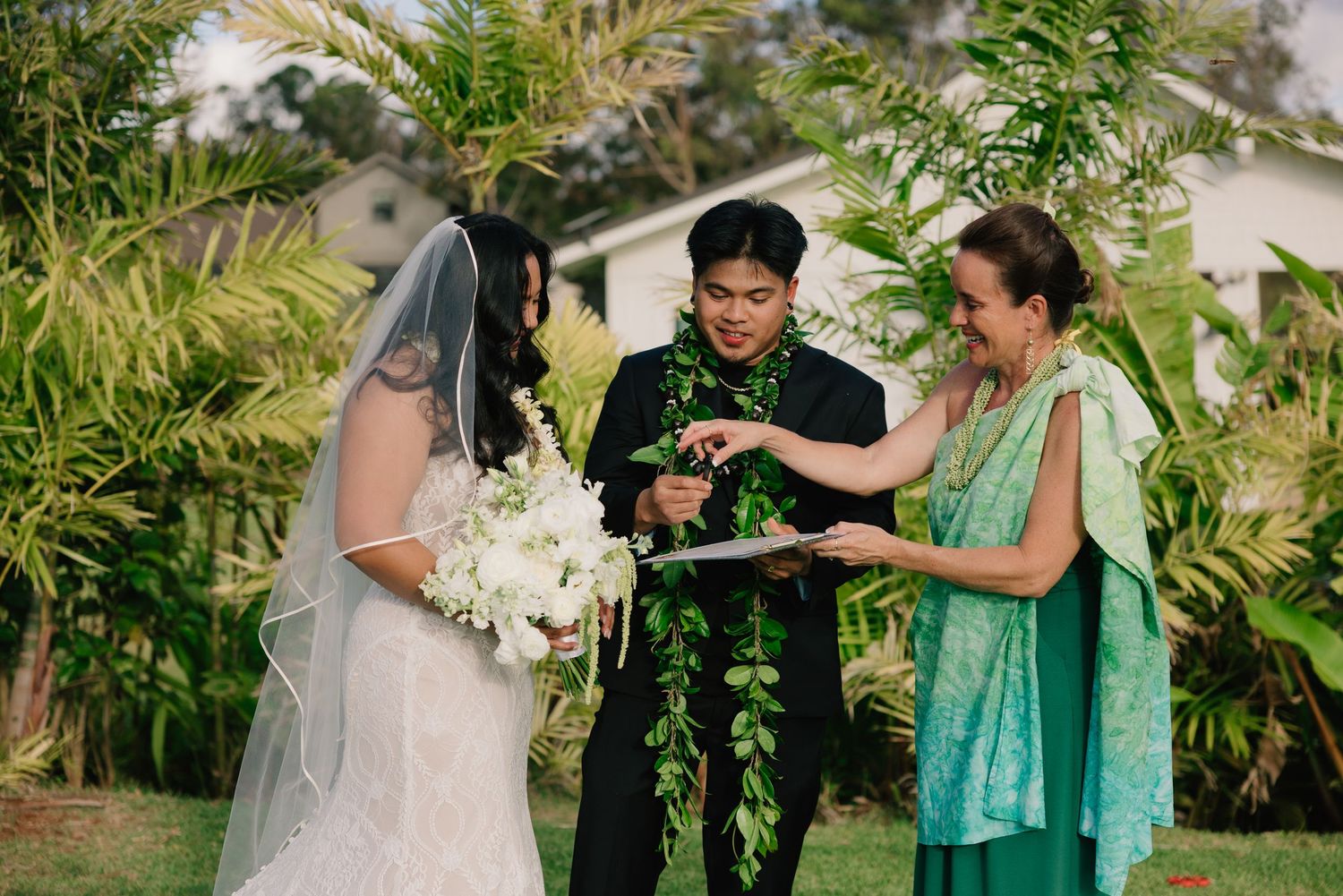 Outdoor Hawaiian wedding ceremony with bride in veil, groom in lei, and officiant in green dress exchanging rings.