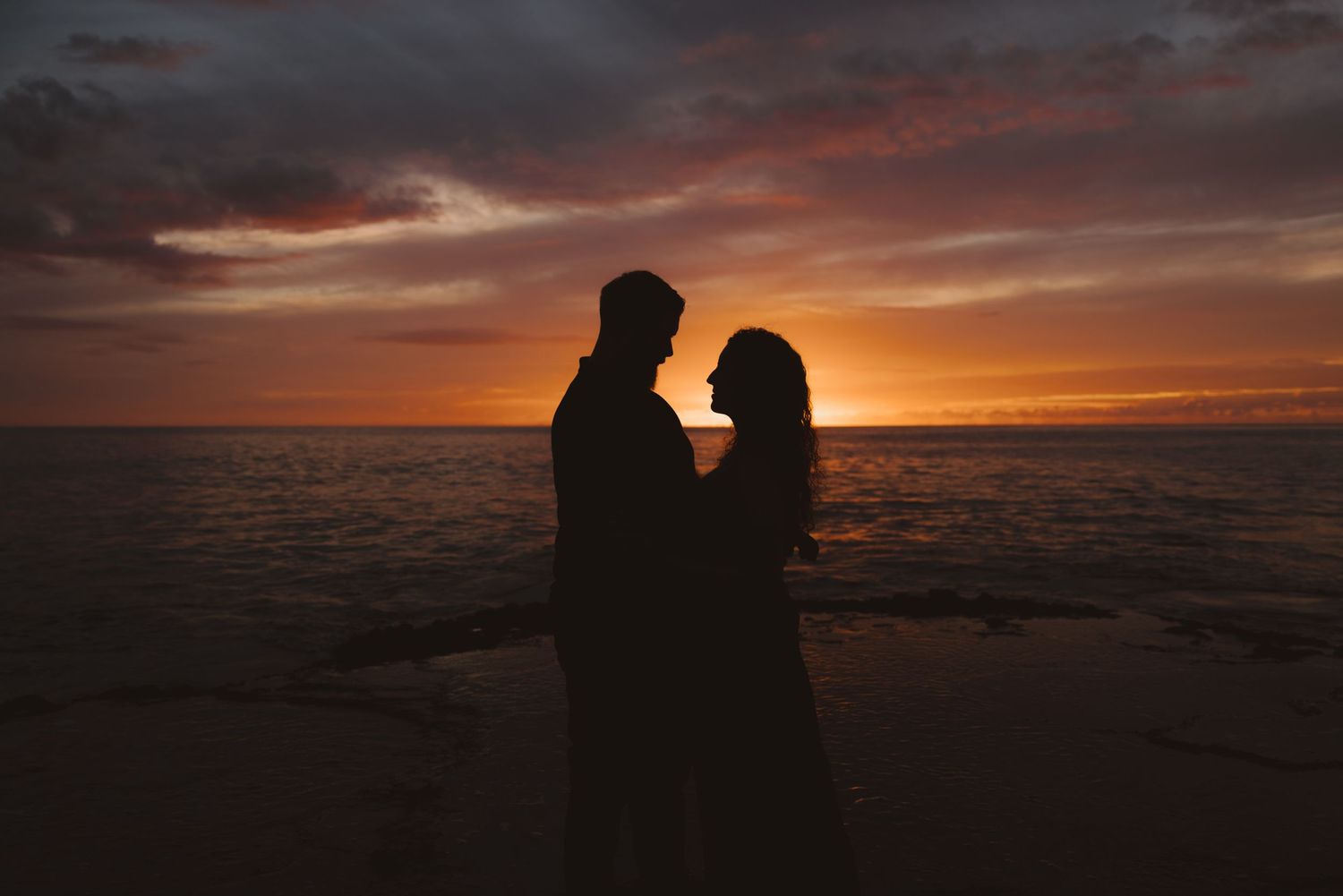 Silhouetted couple warmly embrace against a dramatic orange and purple oahu sunset sky over a vast ocean and beach landscape.