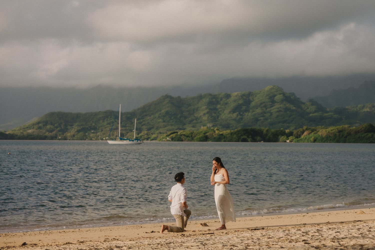 Man proposing on one knee to woman on tropical beach with lush green mountains and sailboat in background.