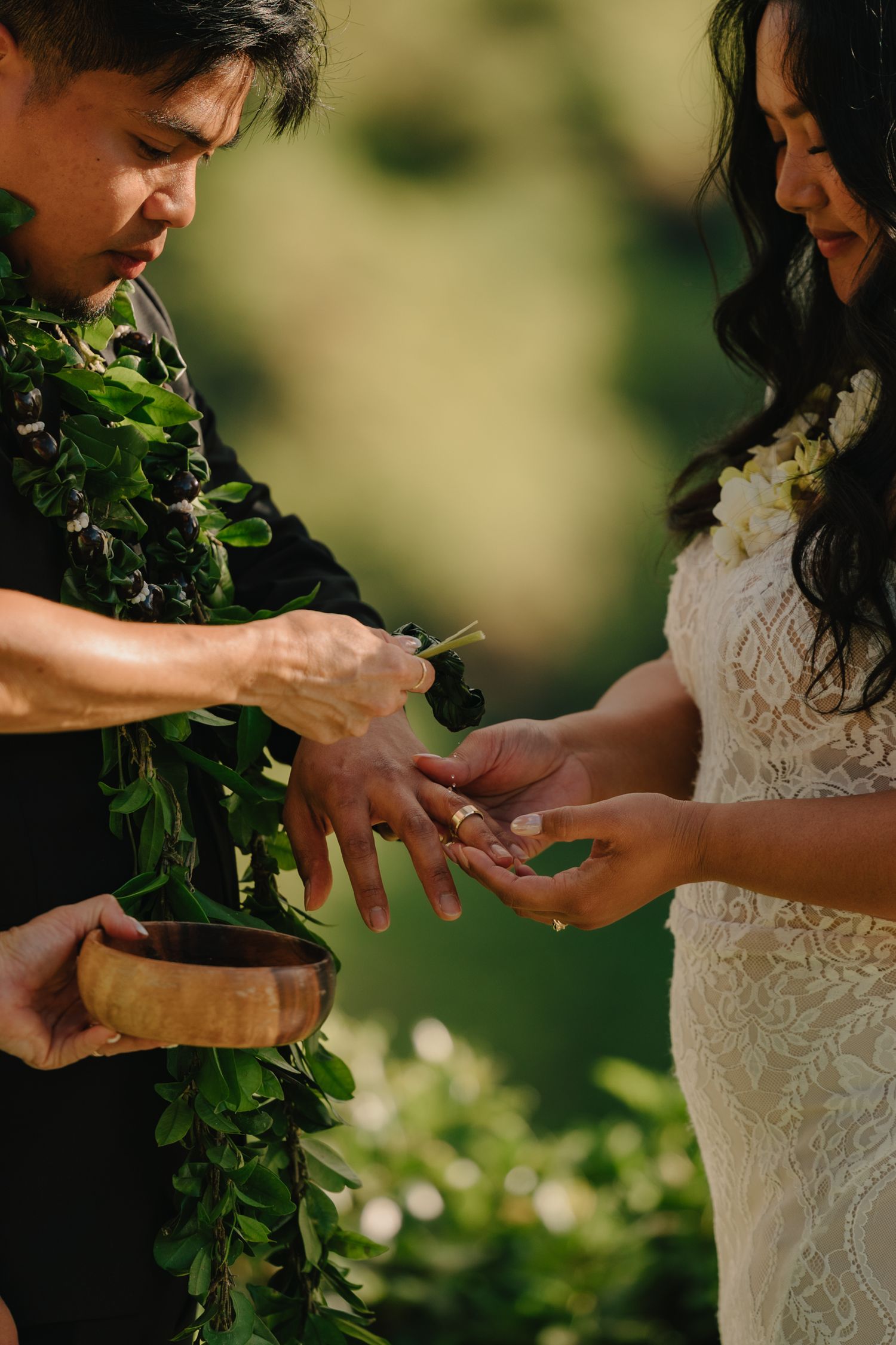 Hawaiian wedding officiant placing ring on bride's hand during outdoor ceremony with green lei and wooden bowl.