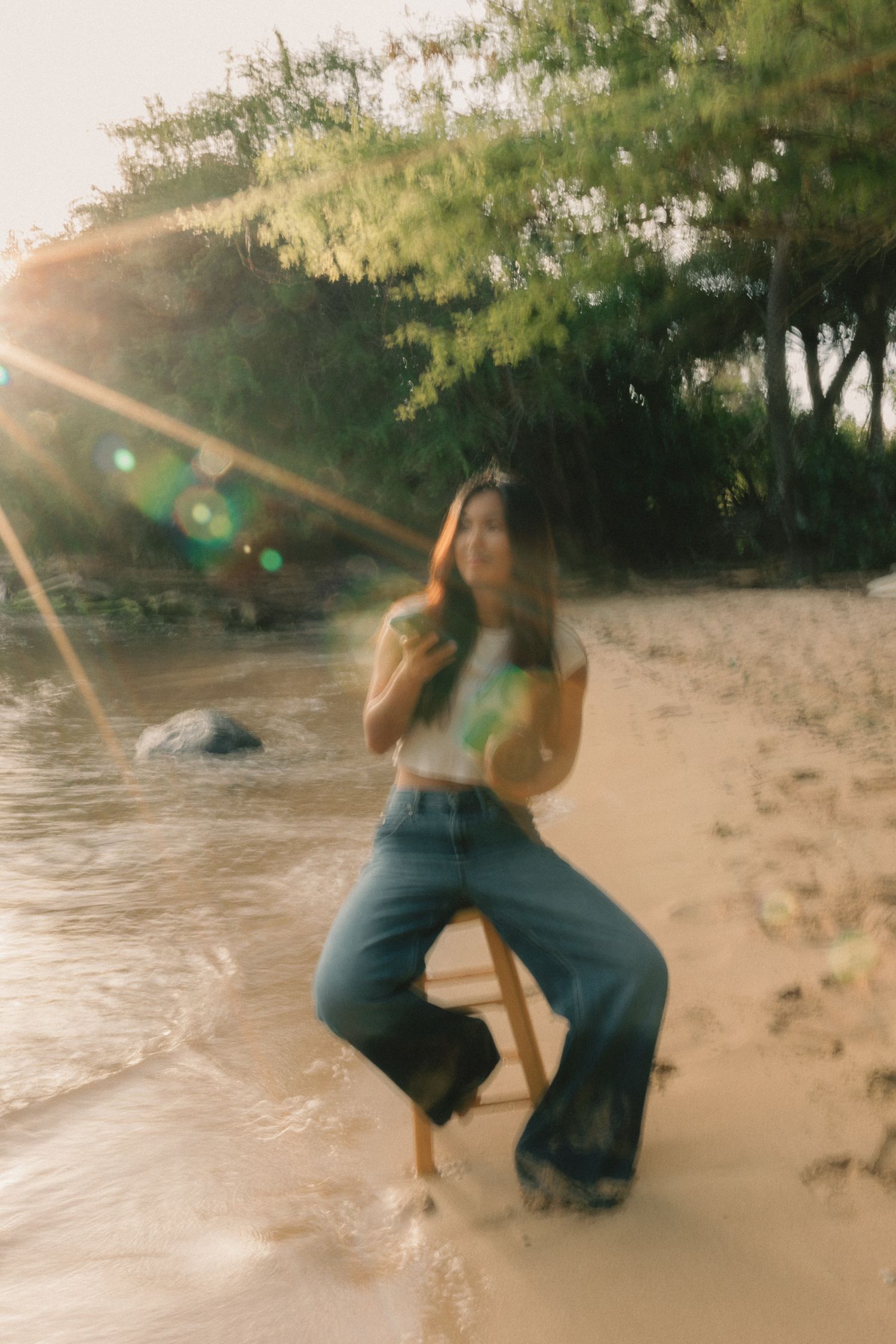 Woman in wide-leg jeans sits on stool at sandy beach with sun flare and lush green trees at golden hour.