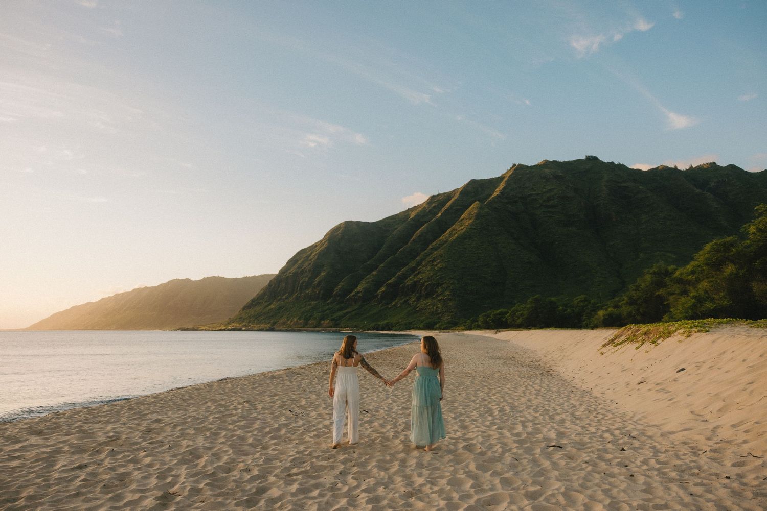 Two women hold hands on a sandy Hawaiian beach at sunset with lush green mountains in the background.