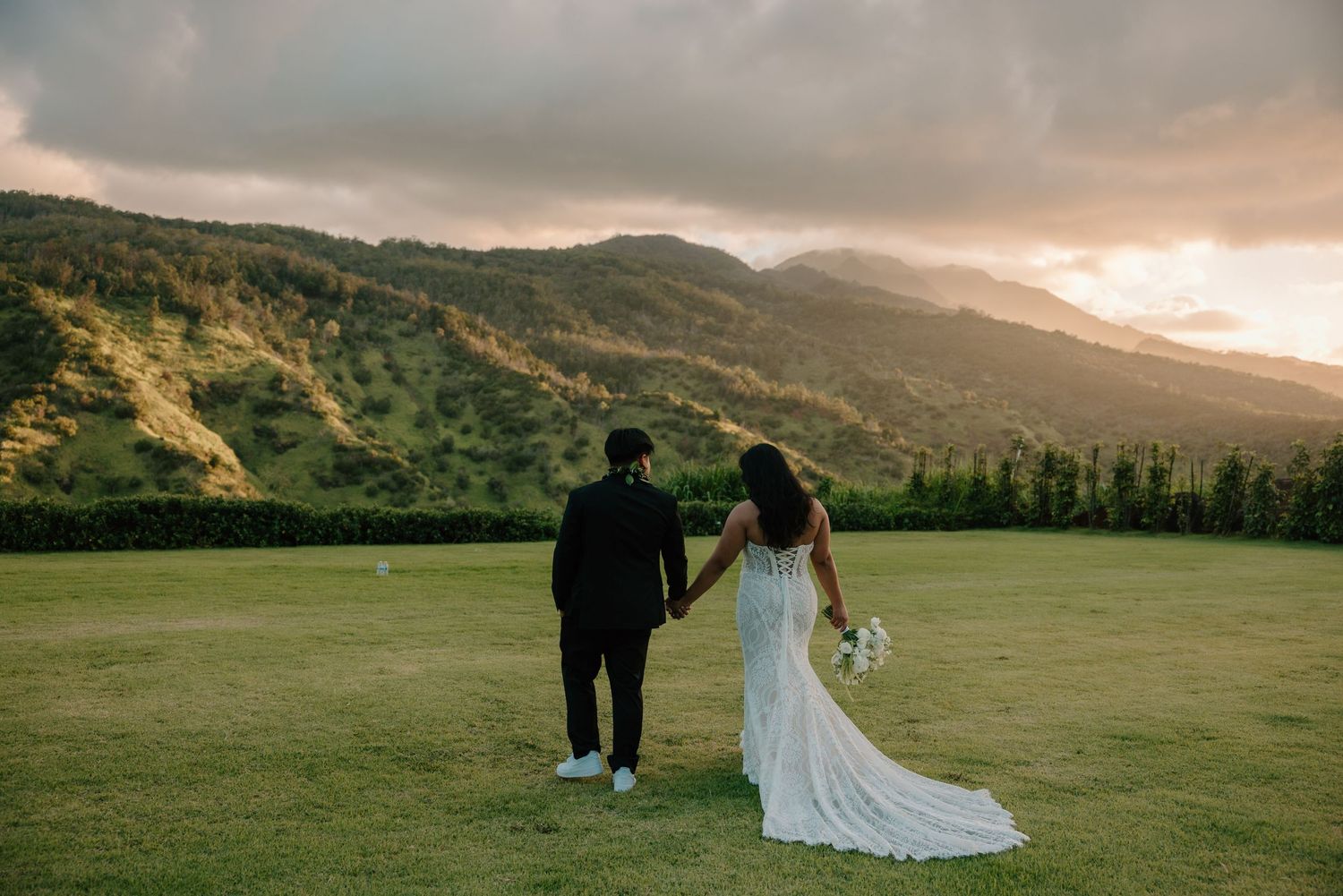 Bride and groom holding hands at sunset overlooking lush green mountains in Hawaii wedding photo.