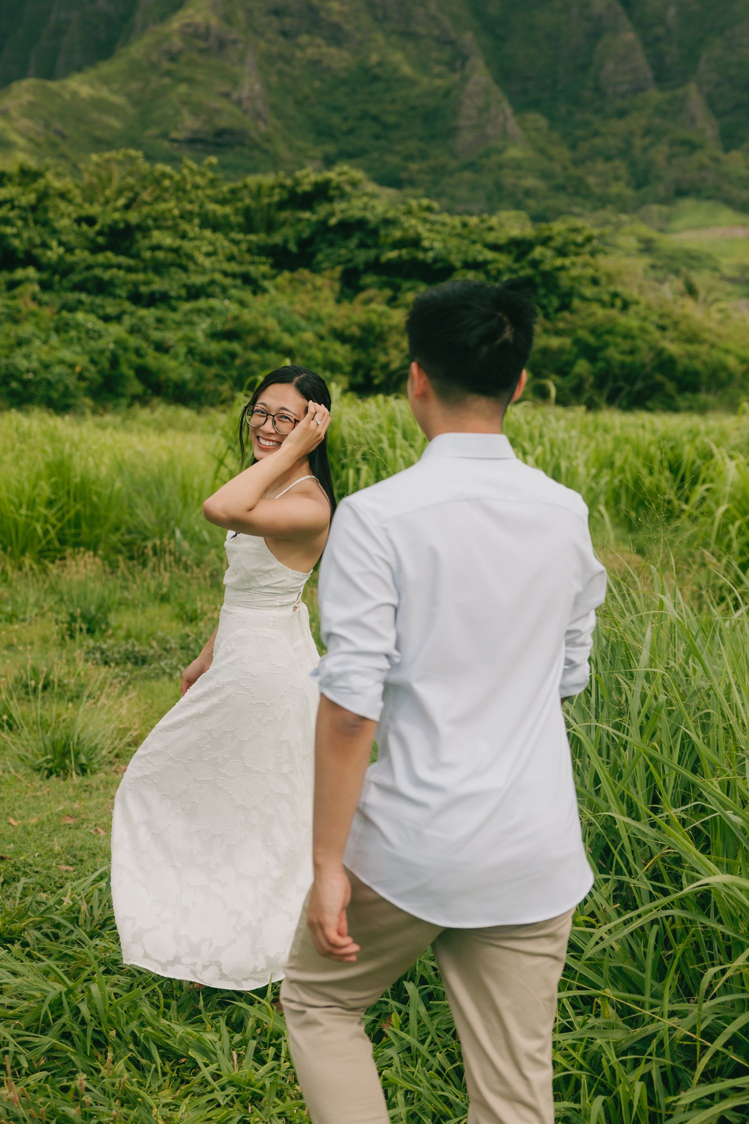 A couple walks through a lush green field, woman in white dress laughing, man in white shirt, with mountains behind.