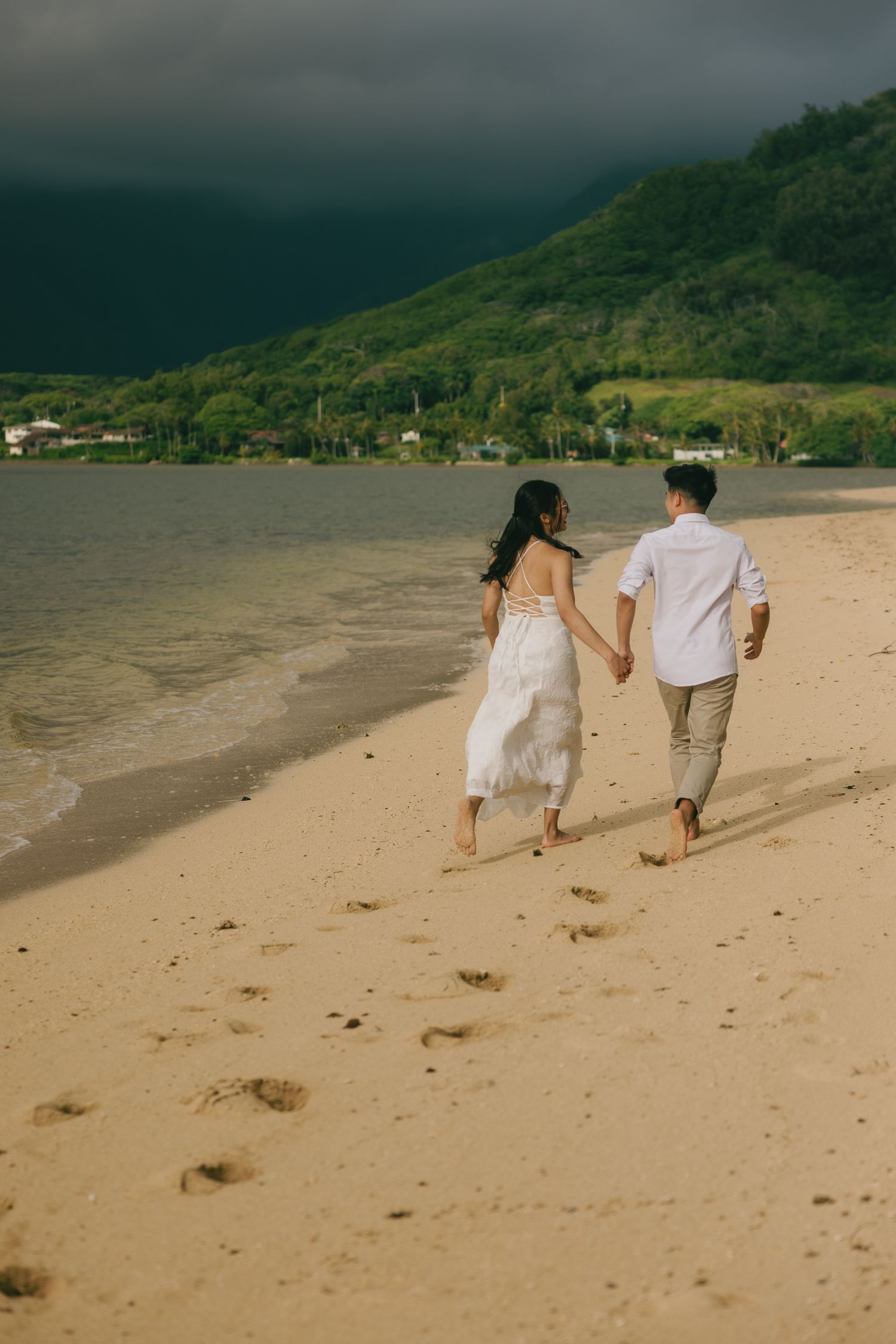 Couple holding hands walking barefoot on sandy beach with green hills and dramatic dark sky in background.