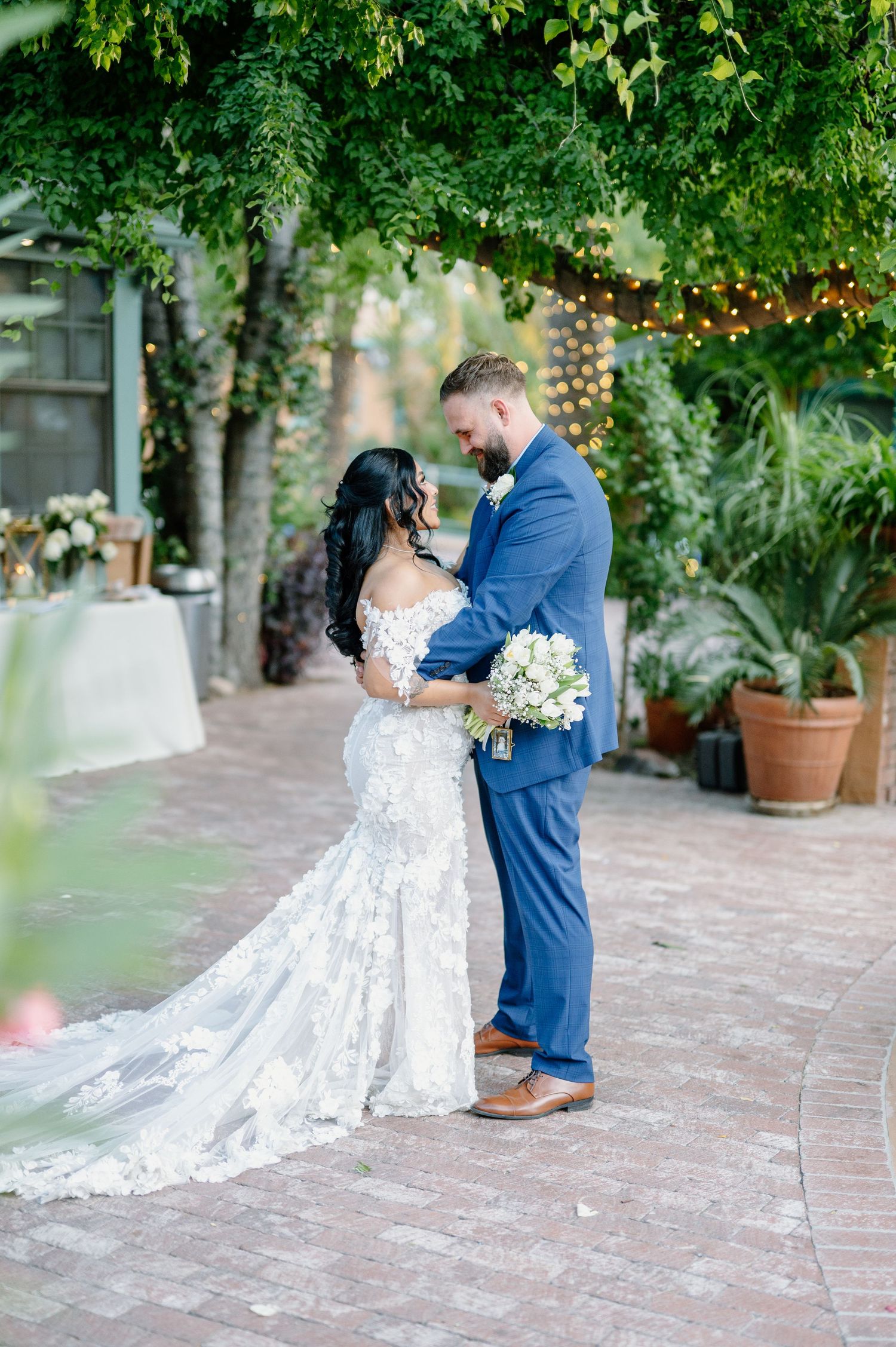 A romantic outdoor courtyard wedding ceremony with string lights and greenery, featuring a lovely lace gown and blue suit.