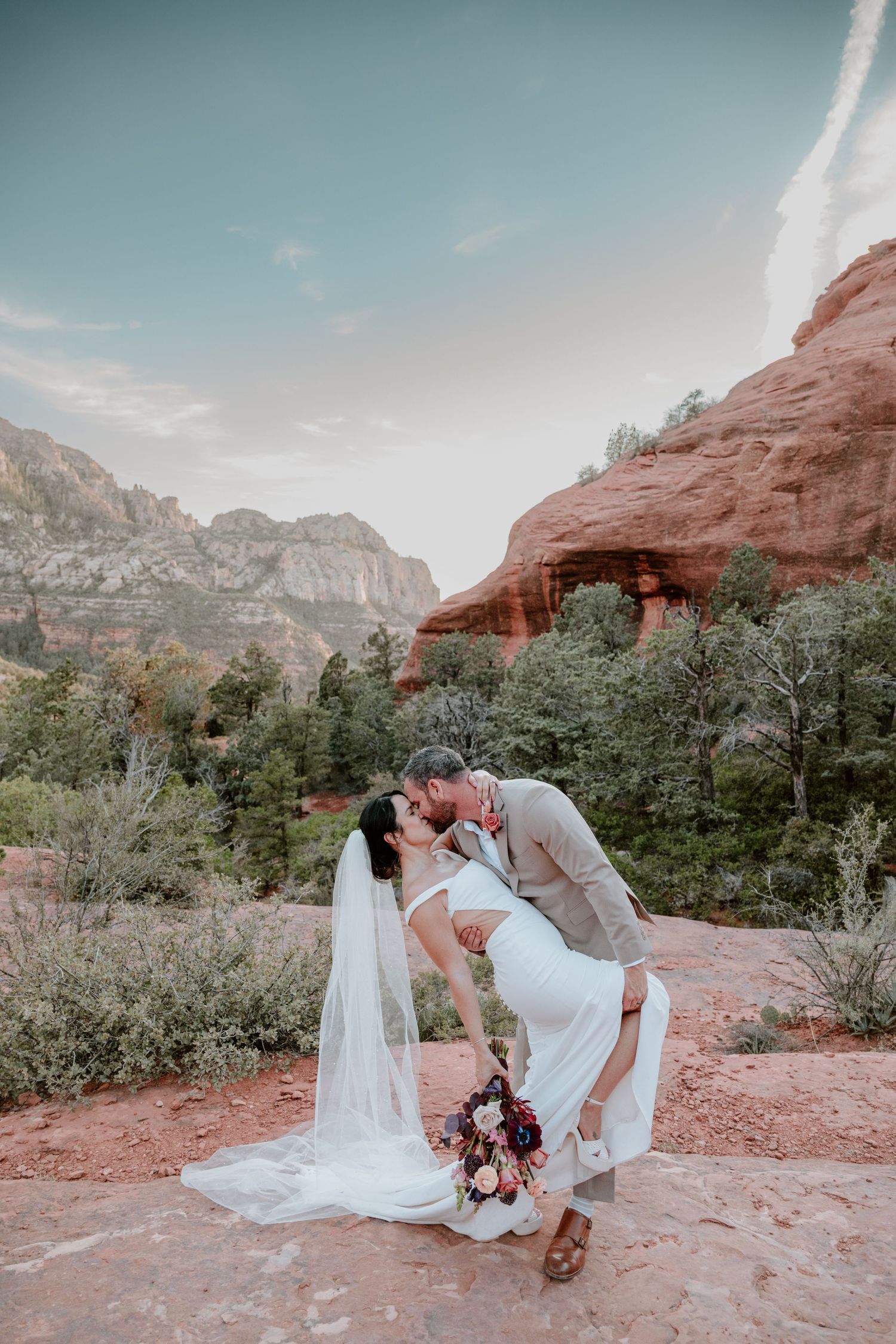 Romantic couple shares a dip kiss against stunning red rock formations and desert landscape at Sedona wedding venue.
