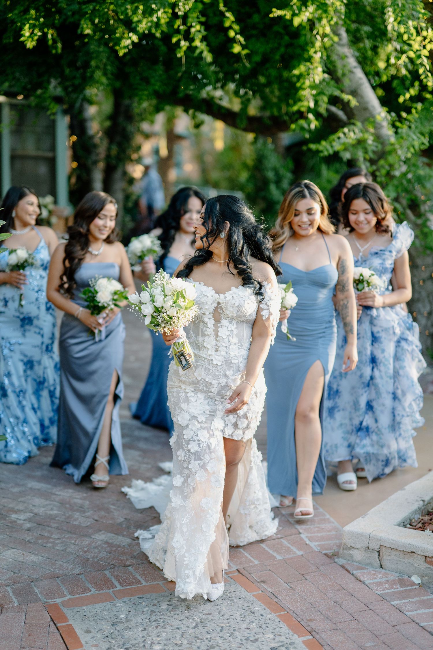 Bridesmaids in flowing dusty blue dresses walk together while holding white floral bouquets during an outdoor wedding.