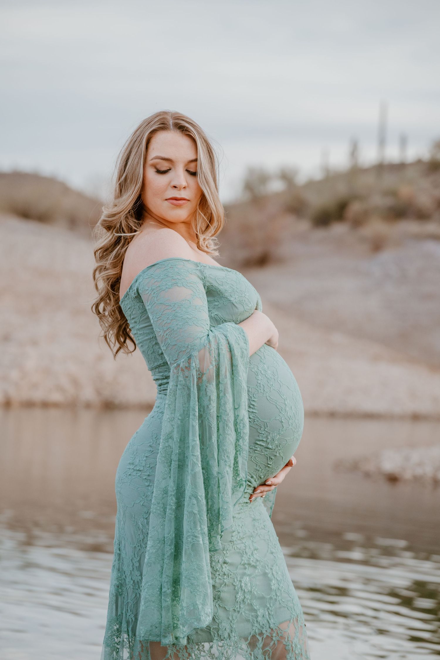 A pregnant woman in a turquoise lace maternity dress poses by the water during a sunset photoshoot.