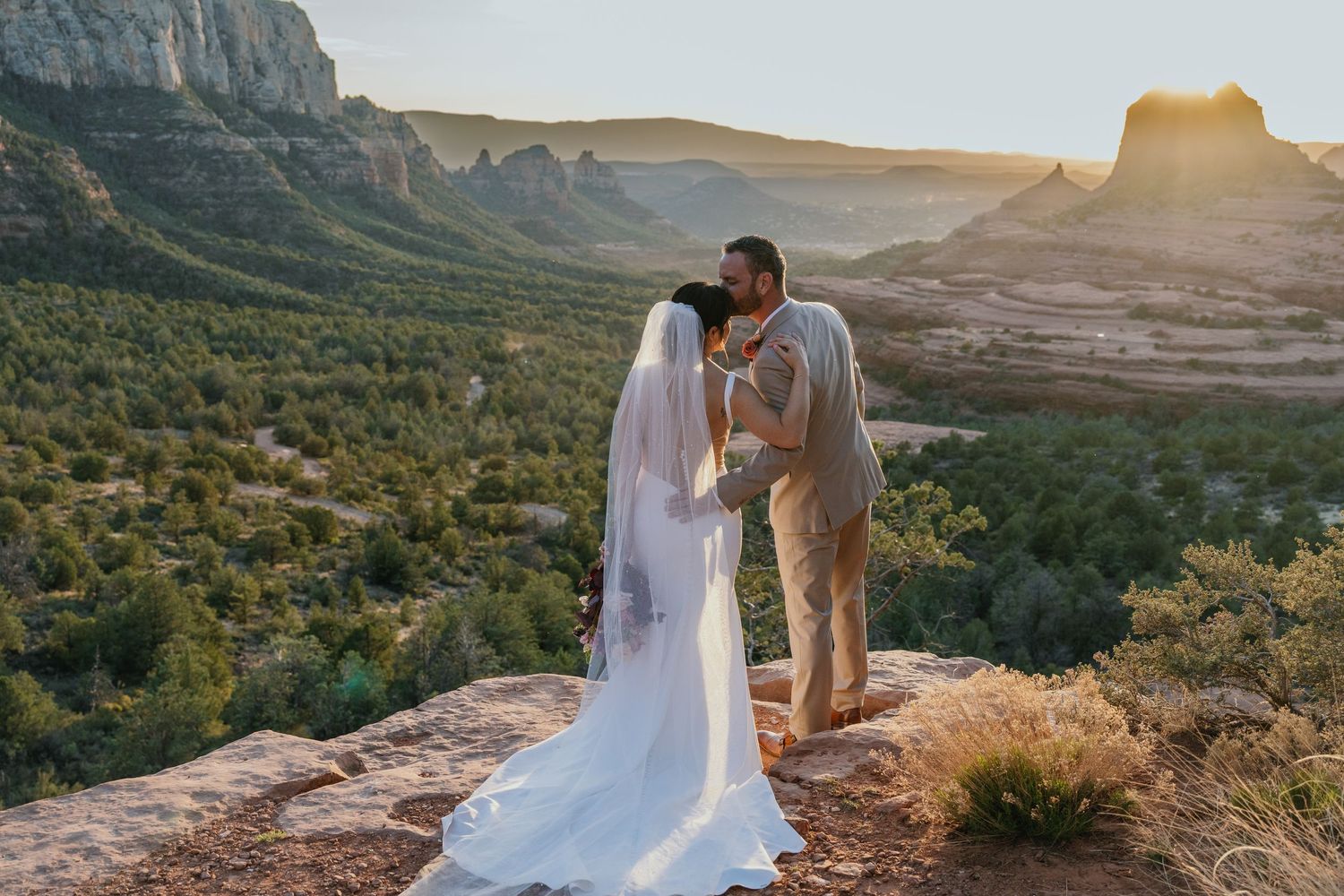 A couple shares a romantic kiss at sunset on a cliff overlooking the desert landscape with mountains in the background.