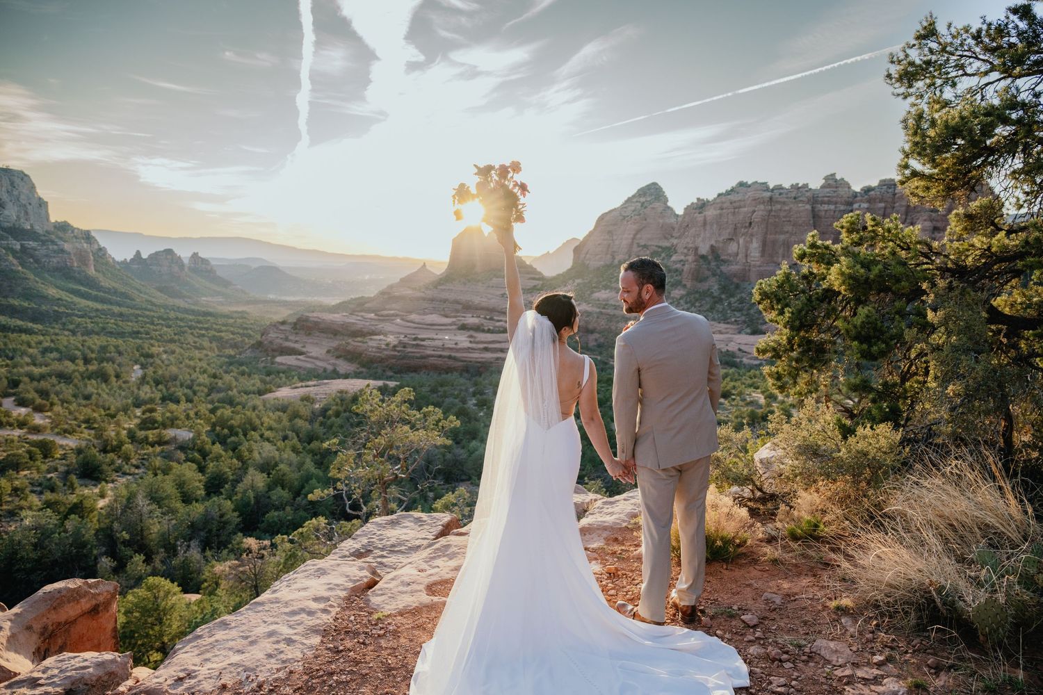 A couple in wedding attire stands on a red rock overlook with scenic mountain vistas in Sedona at sunset.