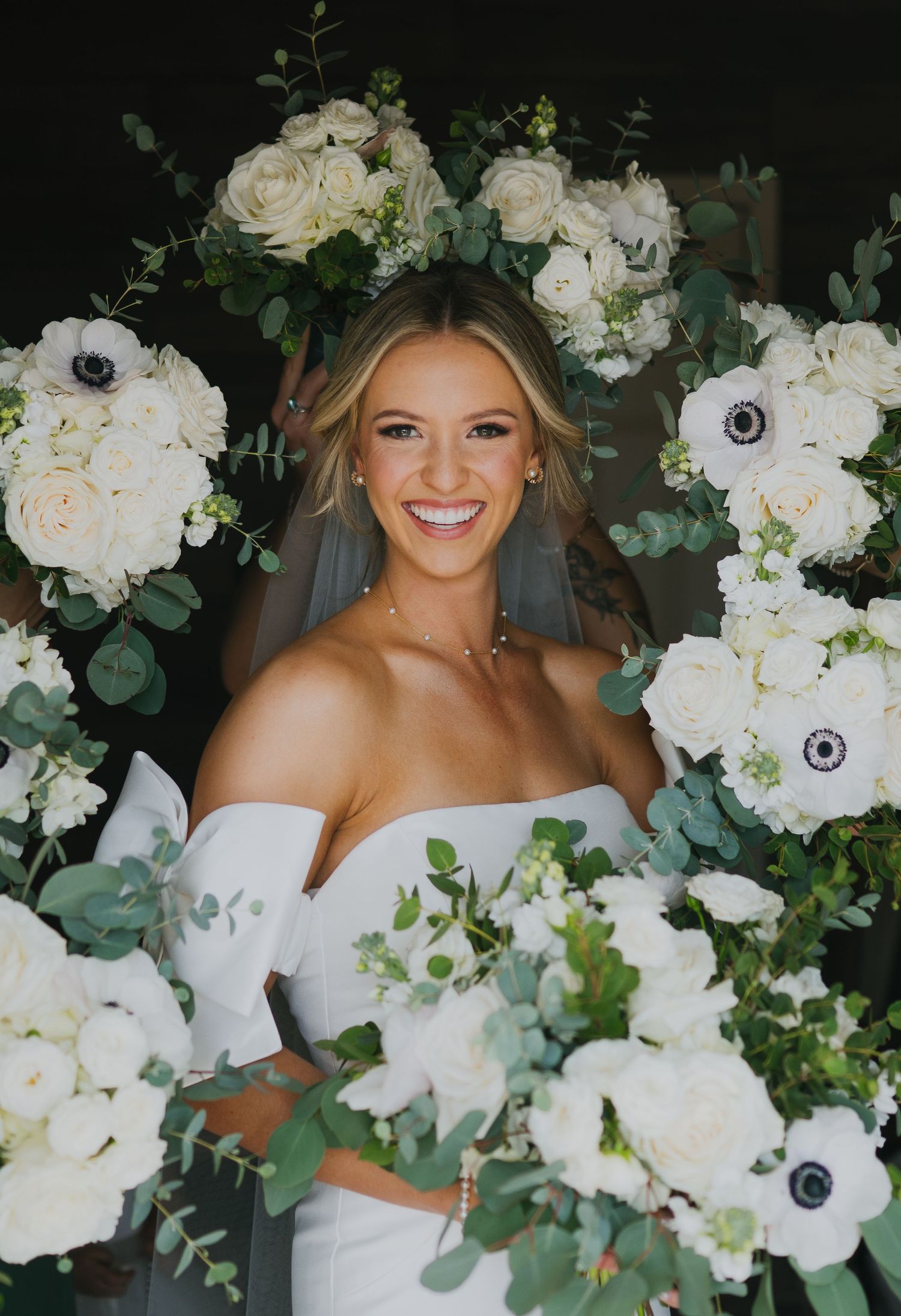 A bride in a strapless white gown poses surrounded by lush white floral arrangements with roses, anemones and greenery.