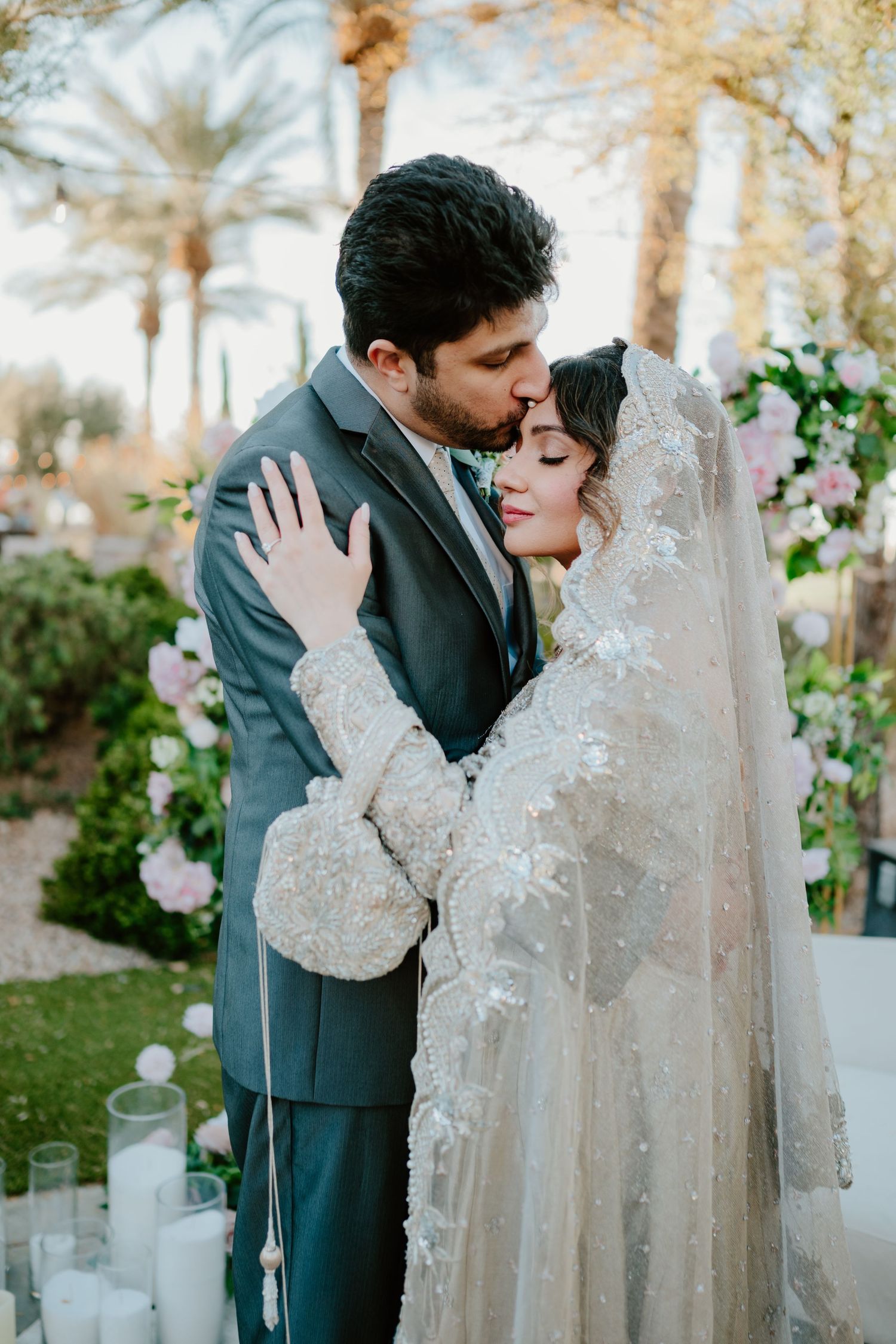 A romantic wedding couple embraces in a garden setting with palm trees and white candles surrounding them.
