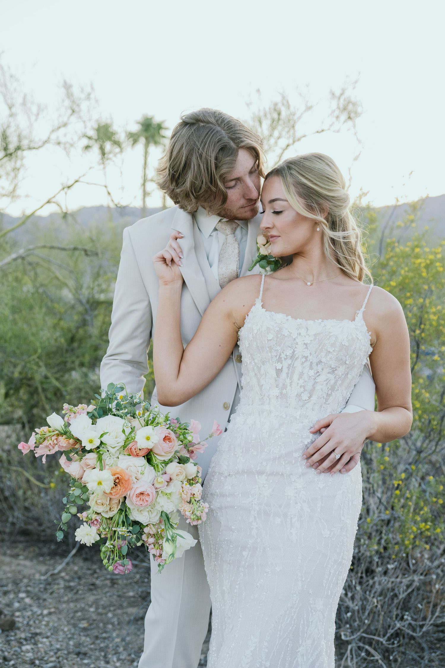 A romantic desert wedding portrait with a white lace dress, light suit, and pastel floral bouquet against mountain scenery.