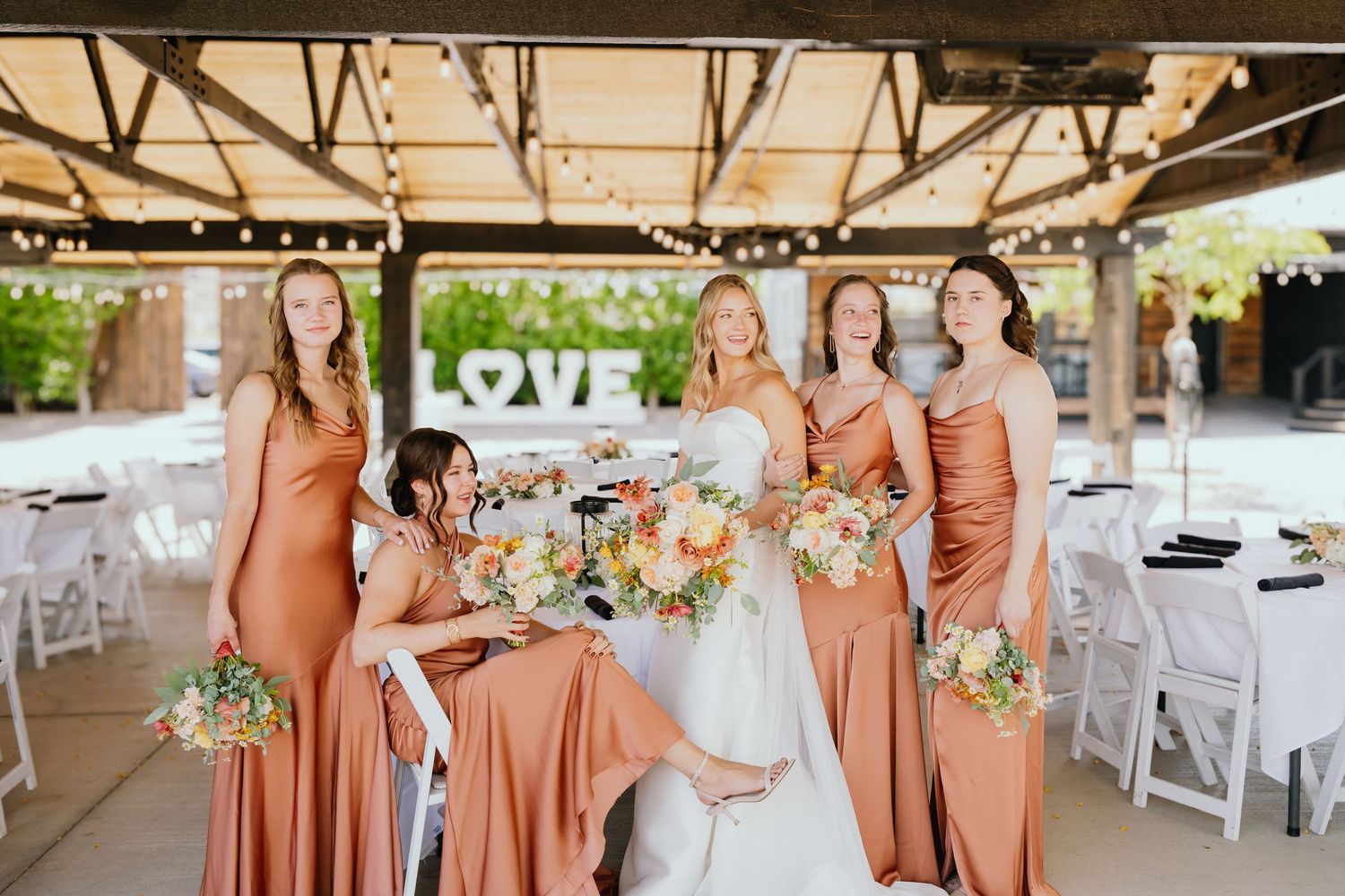 A wedding party wearing peachy-toned dresses poses together at an outdoor venue with floral bouquets.