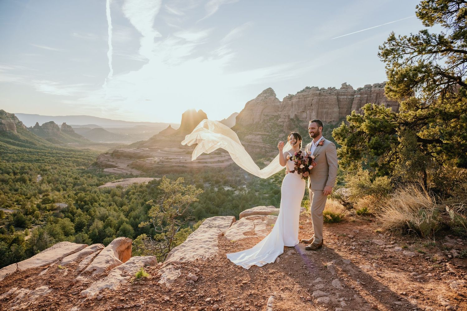 Couple stands on red rock overlook in Sedona with flowing white wedding dress captured during golden hour sunset.