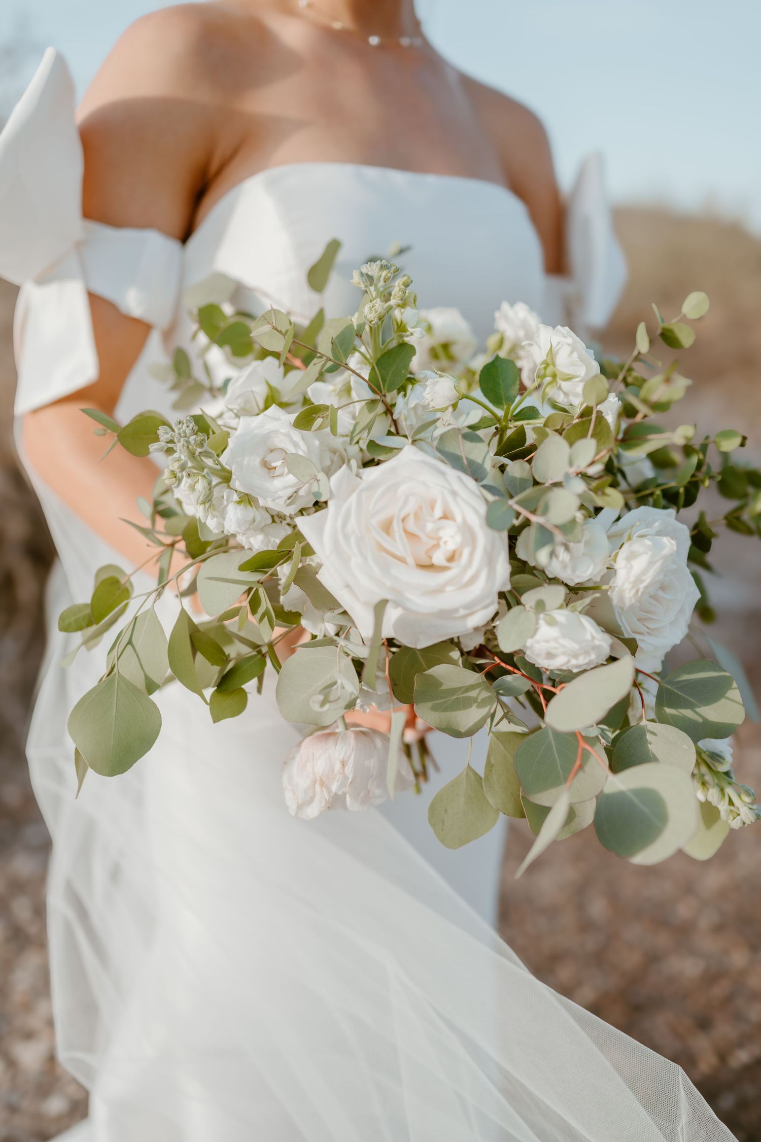 A delicate white wedding bouquet with roses and eucalyptus held against an ivory wedding dress in soft natural light.