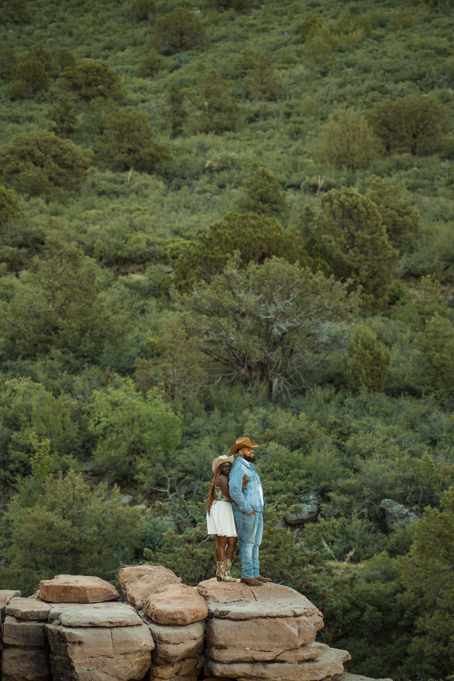 Two figures stand on rocky cliffs surrounded by lush green forest landscape during a nature photoshoot.