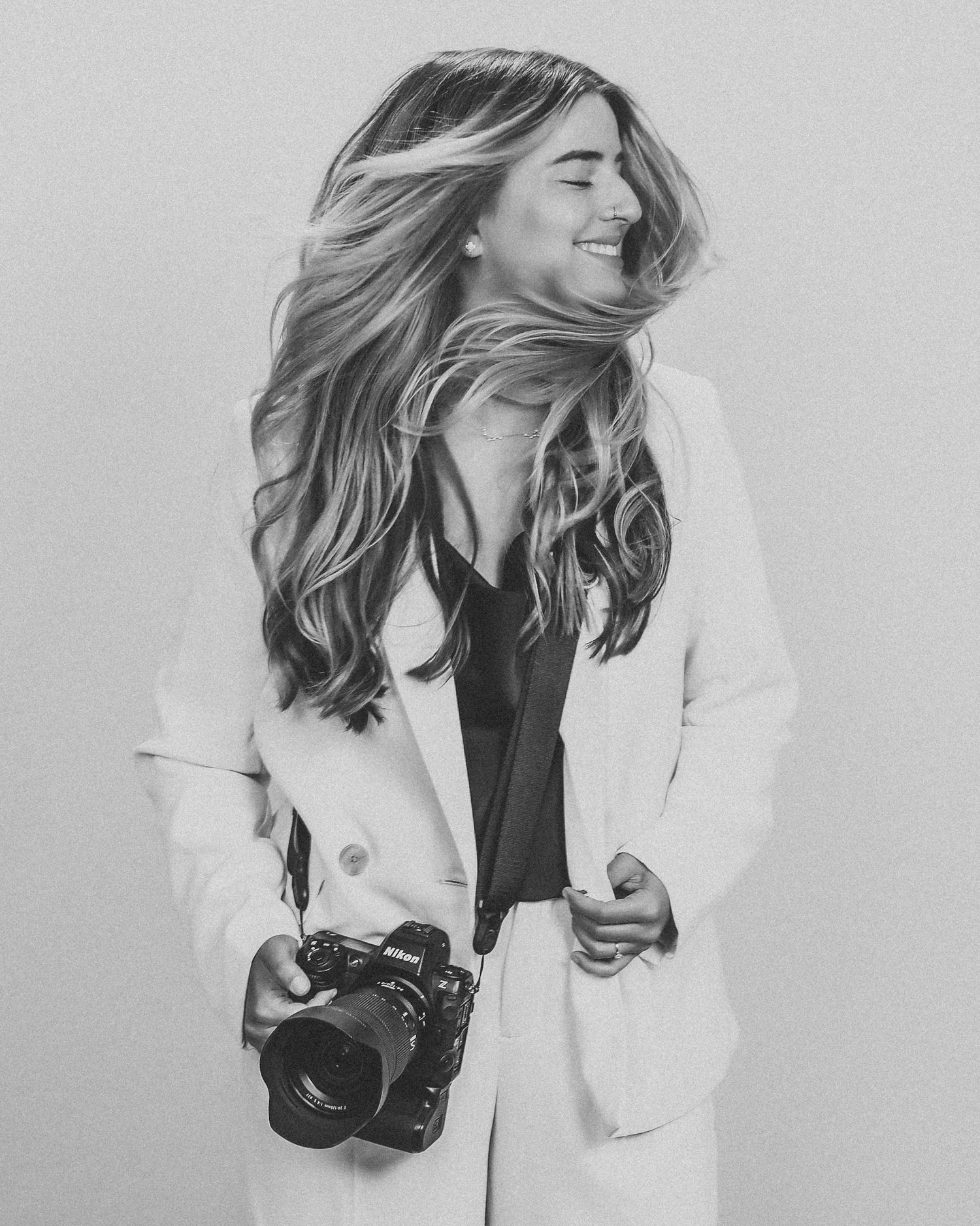 A joyful photographer in a white blazer and black top laughs while holding a professional camera against a gray backdrop.