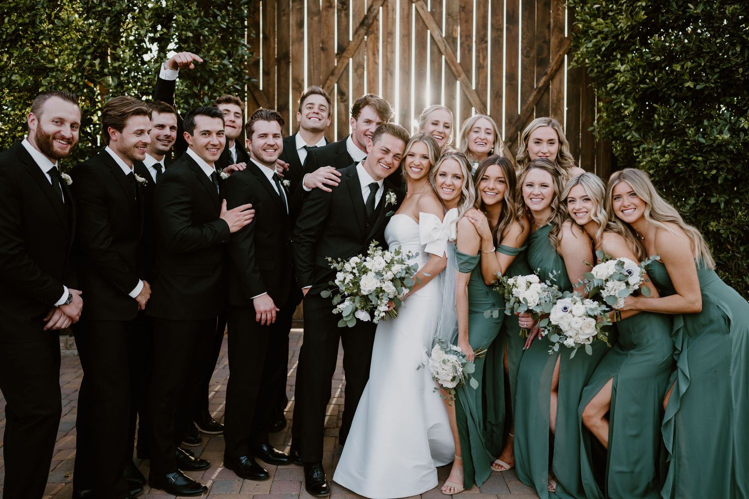 Wedding party poses together with bridesmaids in emerald green dresses and groomsmen in black suits outdoors.