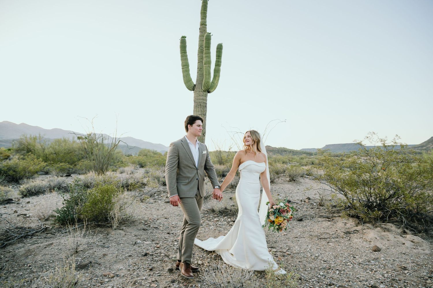 A newlywed couple walks together in front of a towering saguaro cactus in the Arizona desert landscape.