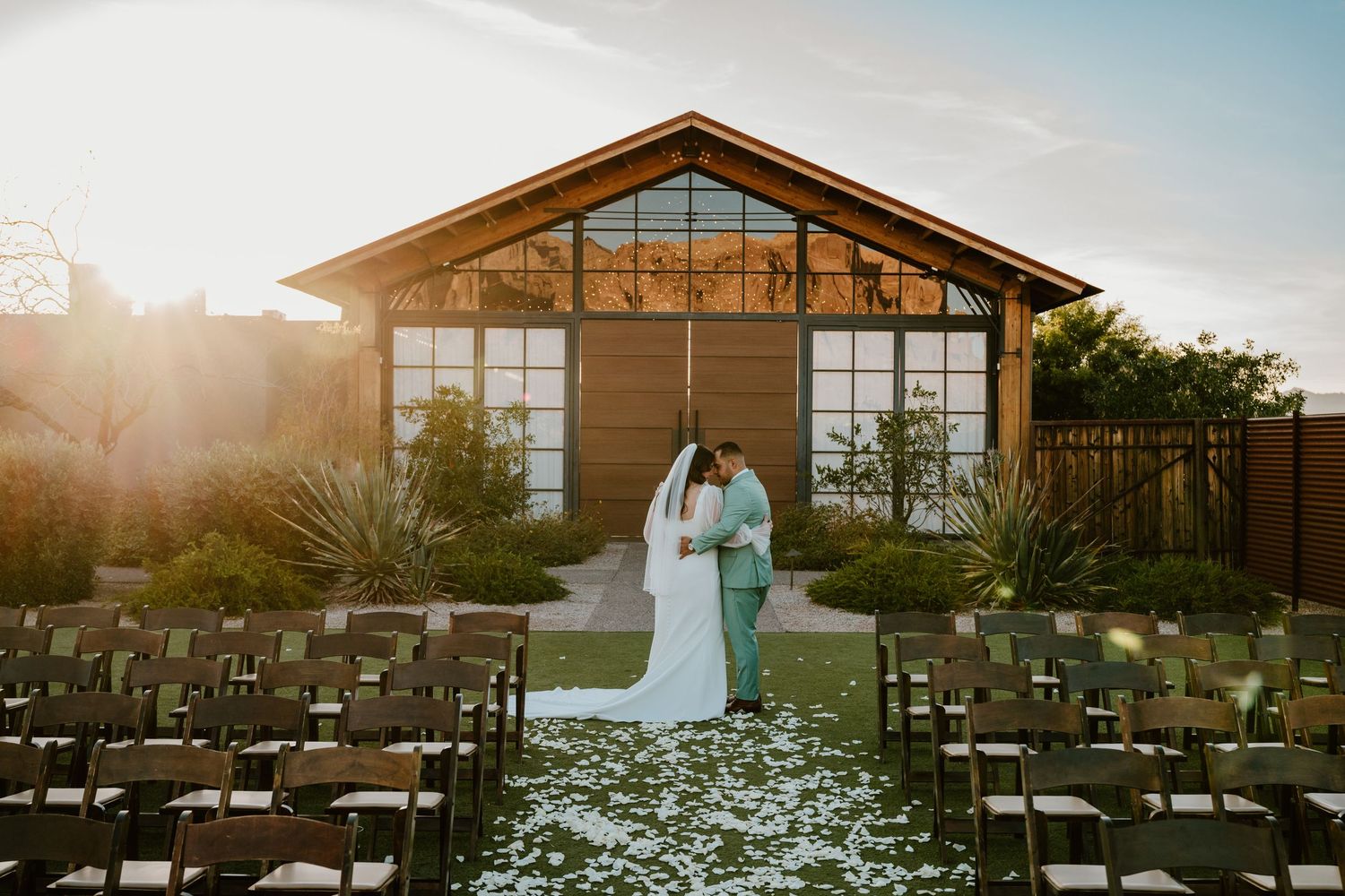 Wedding ceremony setup outside a modern glass-walled venue at sunset with rose petals lining the aisle.