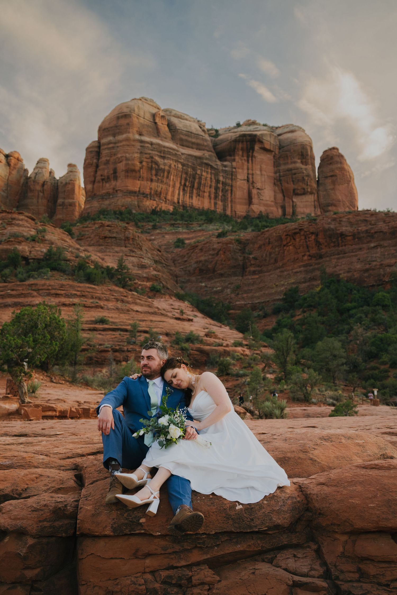 Couple in wedding attire sitting on red rock formations at sunset in Sedona with majestic desert cliffs in background.