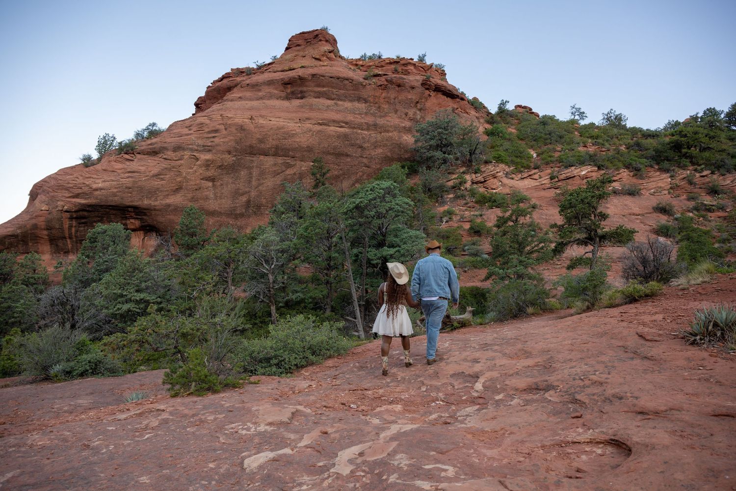 Two hikers explore red sandstone formations along a desert trail in Sedona surrounded by green vegetation.