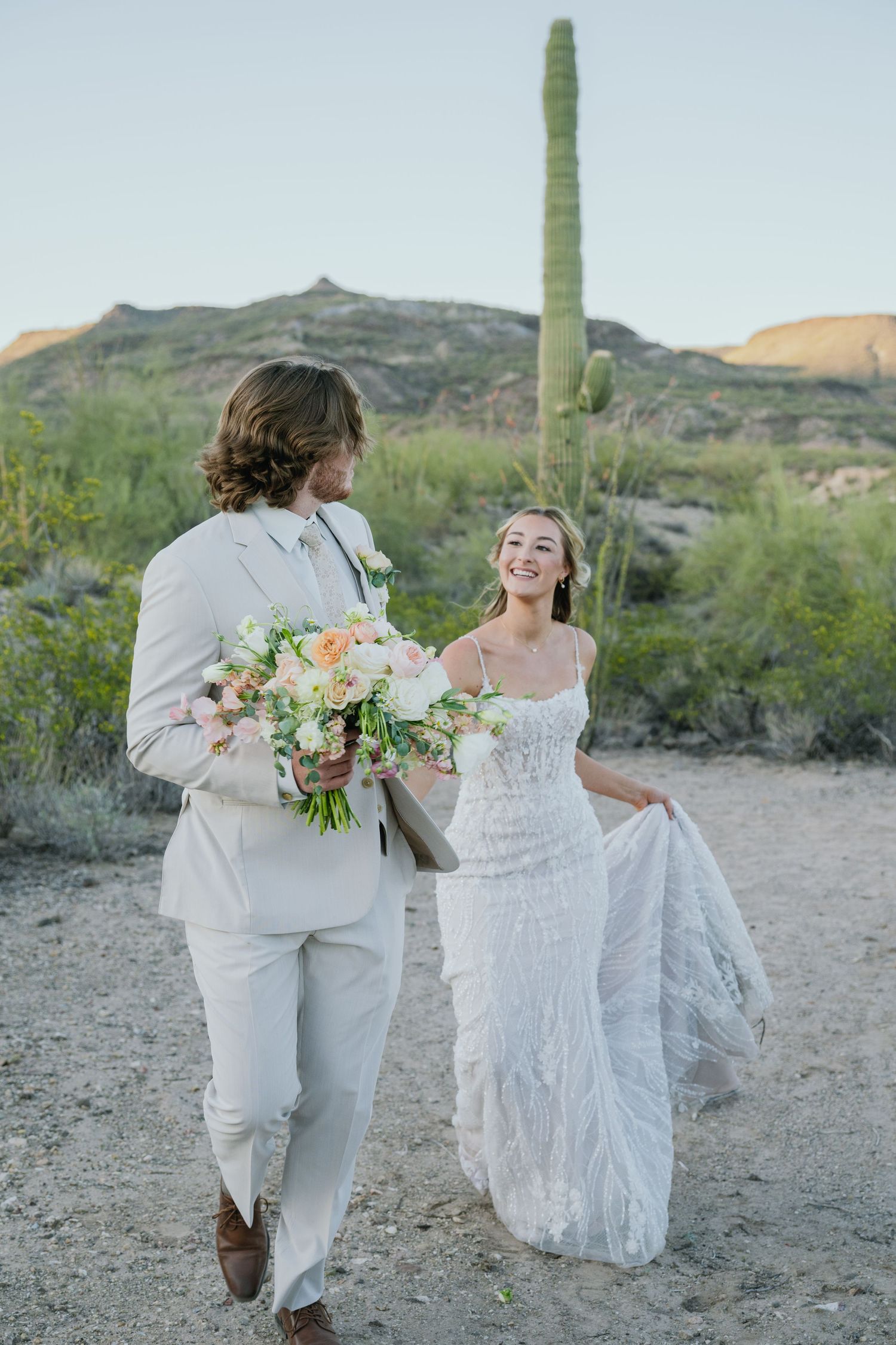 A couple walking together in a desert landscape with saguaro cactus, dressed in wedding attire with flowers.