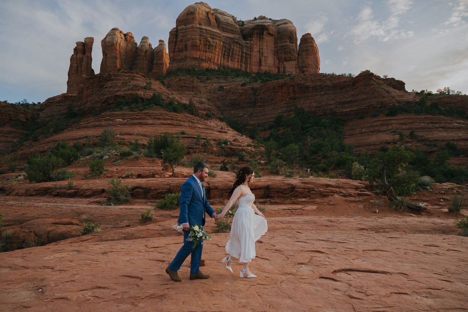 Two people hold hands while walking on red rocks with Cathedral Rock formation in Sedona, Arizona looming behind them.
