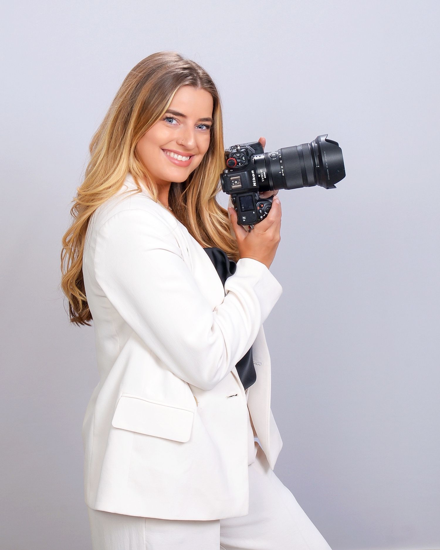 Professional photographer wearing white blazer holds camera while posing against grey background.