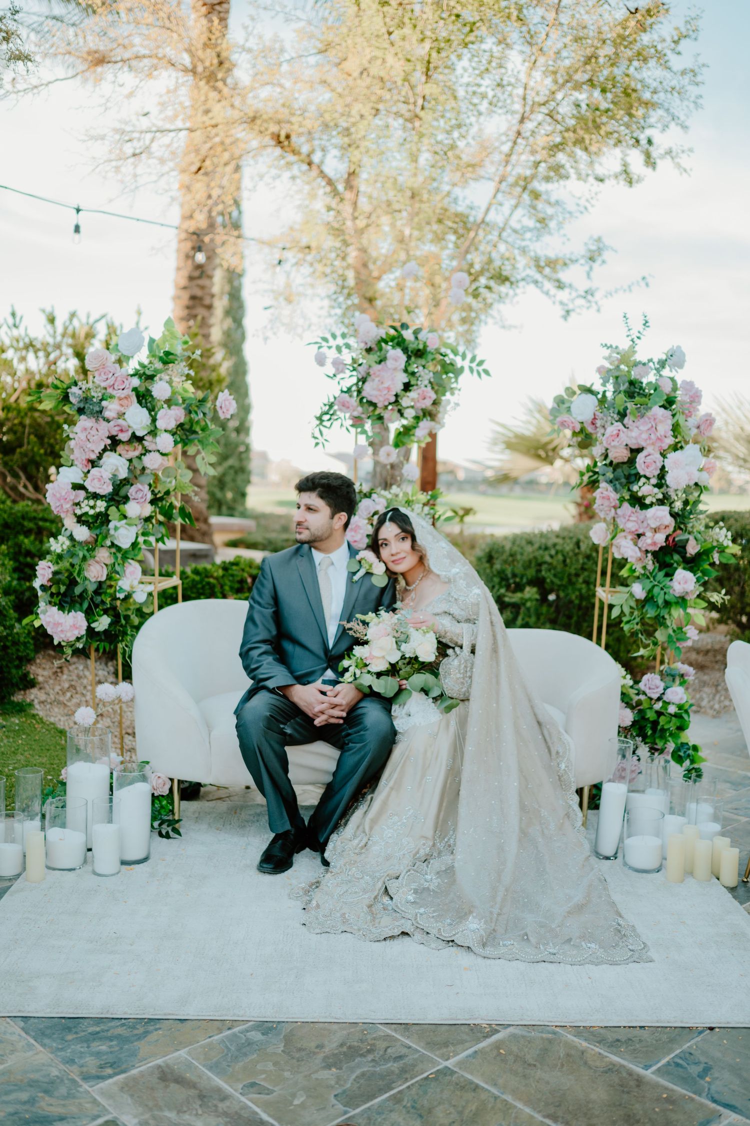 Elegant outdoor wedding setup with floral arches, white candles, and couple seated on white loveseat during sunset.