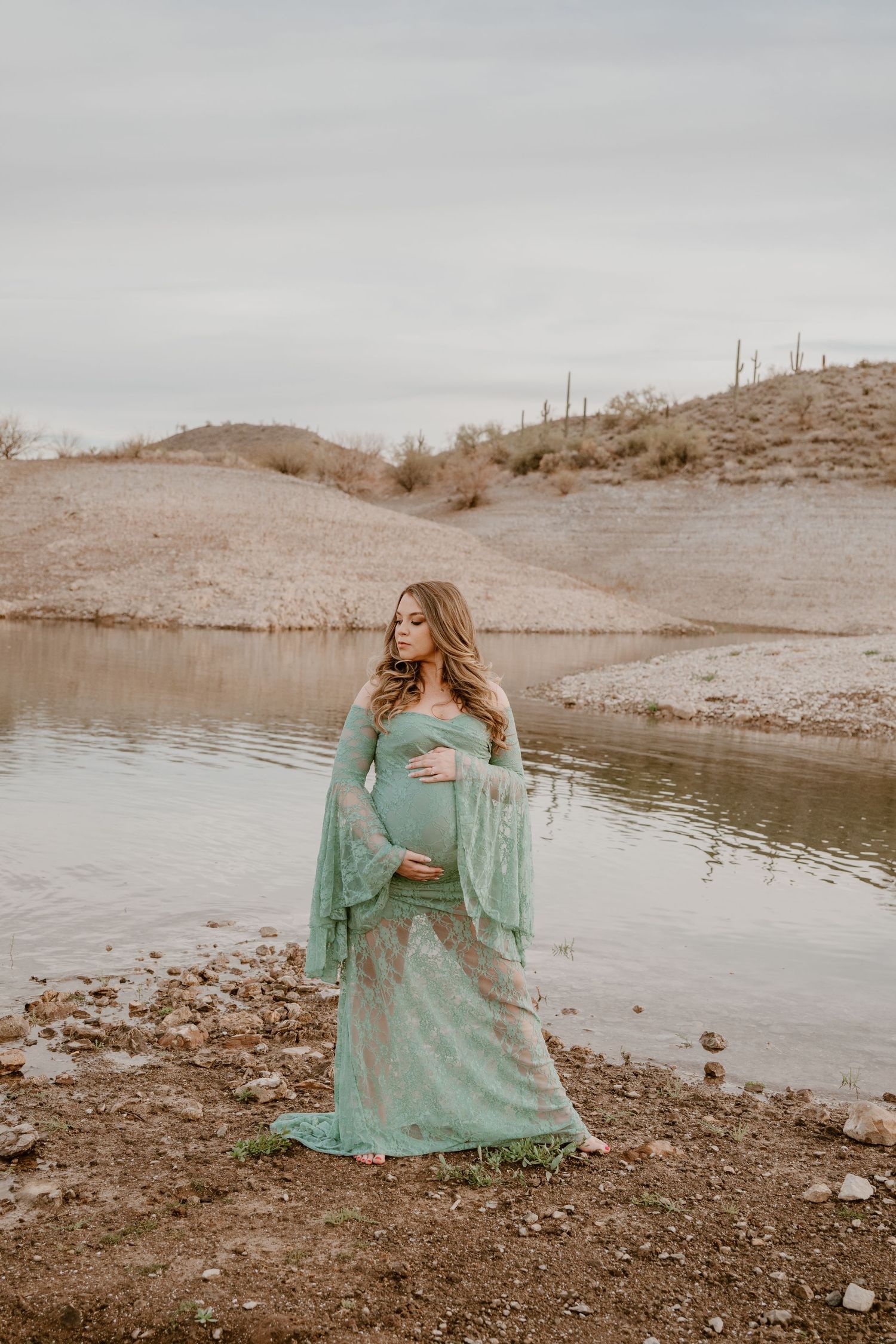 Person in flowing mint green lace dress stands by lake shore in desert landscape during golden hour photoshoot.