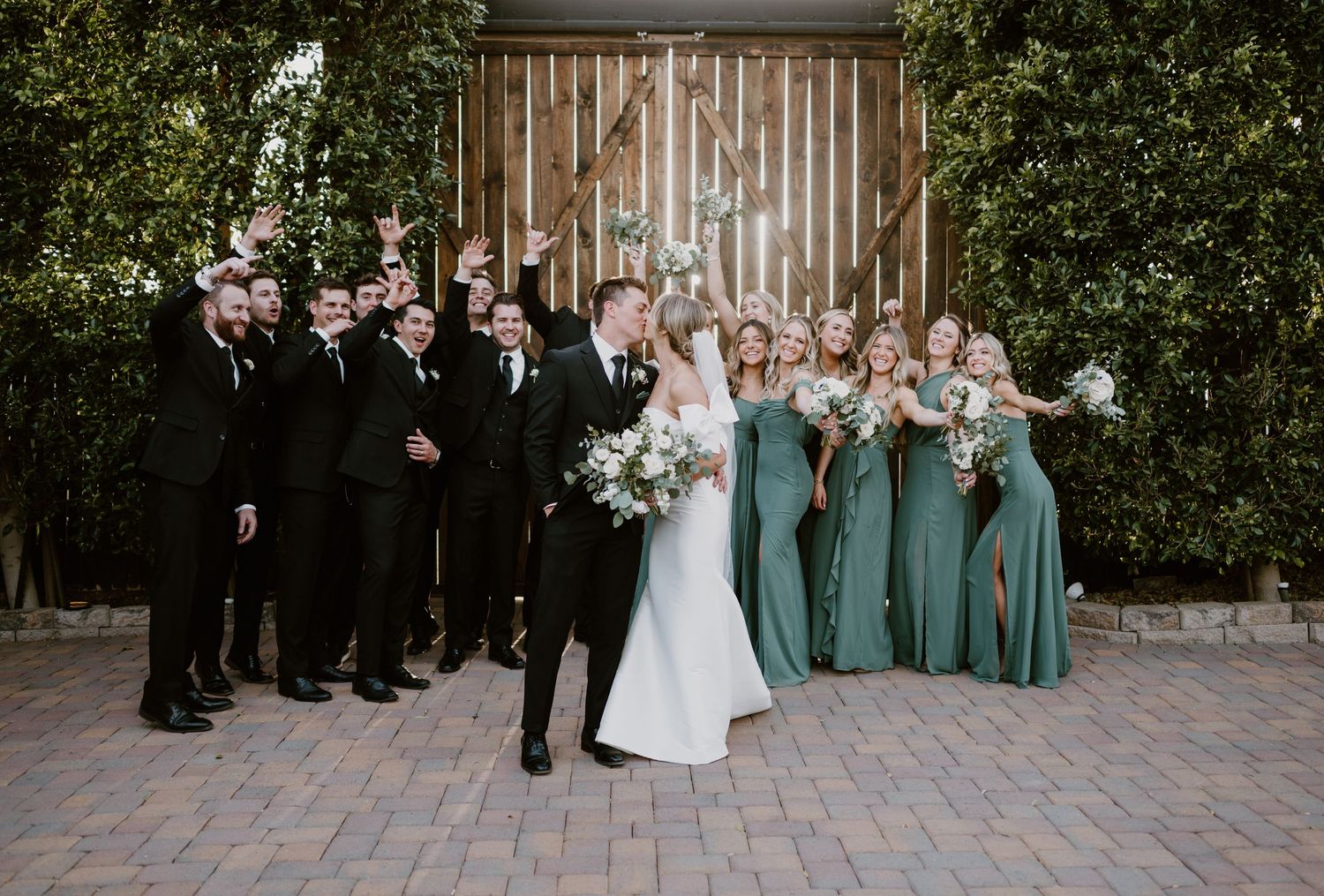 Wedding party in formal attire with bridesmaids in sage green dresses and groomsmen in black tuxedos pose outside rustic venue.