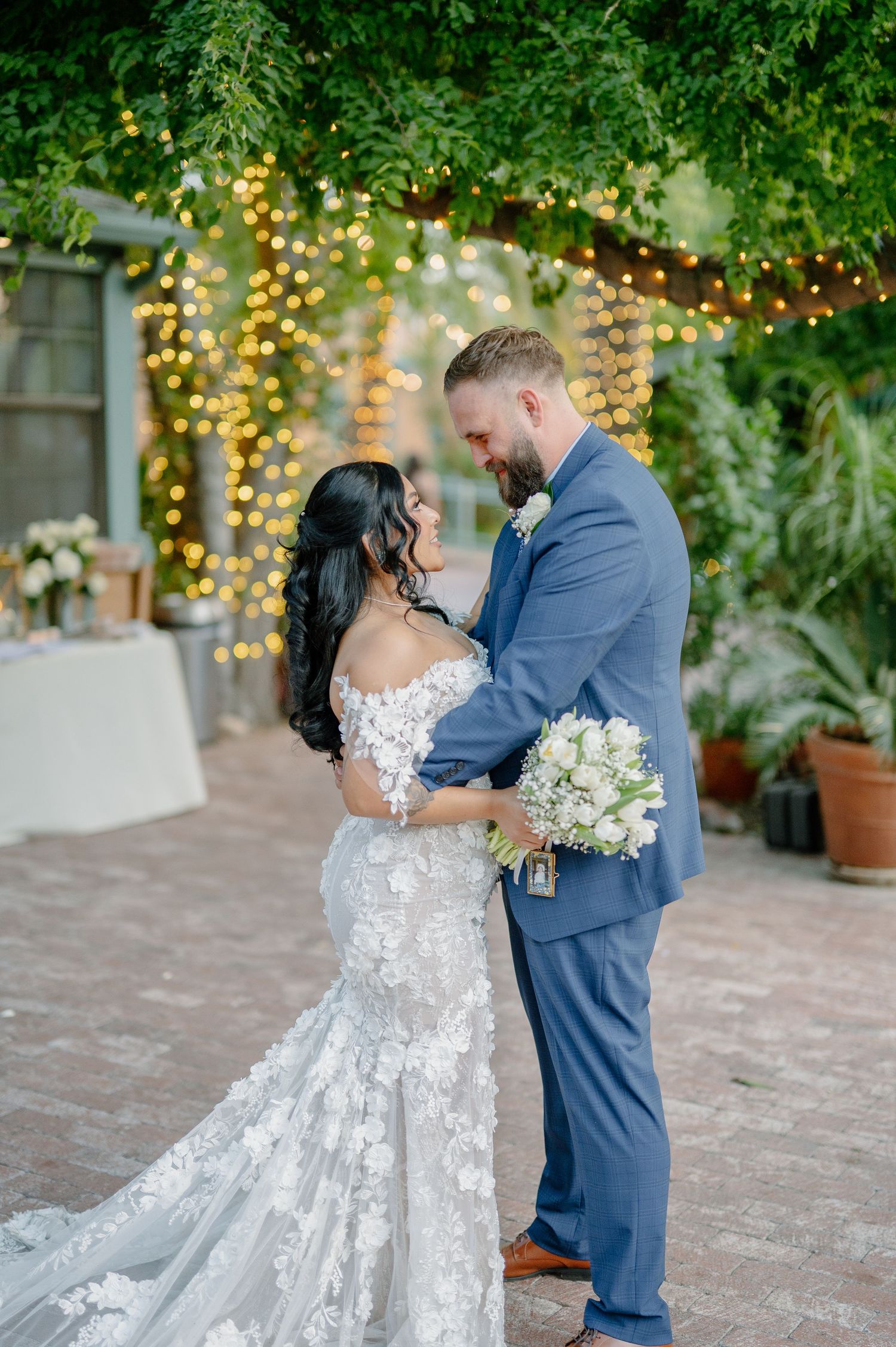 Newlywed couple embraces outdoors under glowing string lights with romantic garden backdrop and floral bouquet.