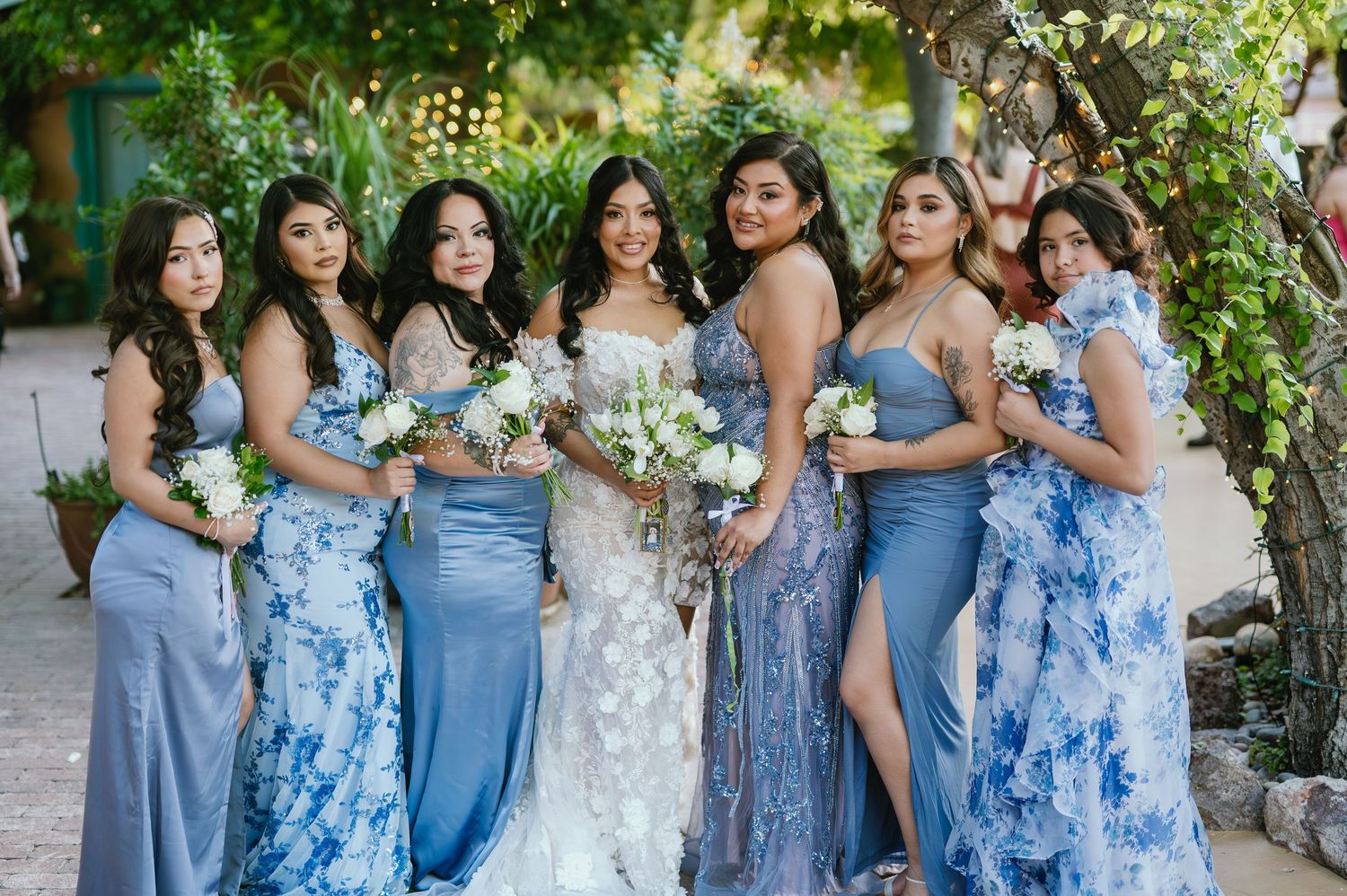A bridal party wearing coordinated blue and floral dresses poses together during an outdoor wedding celebration.