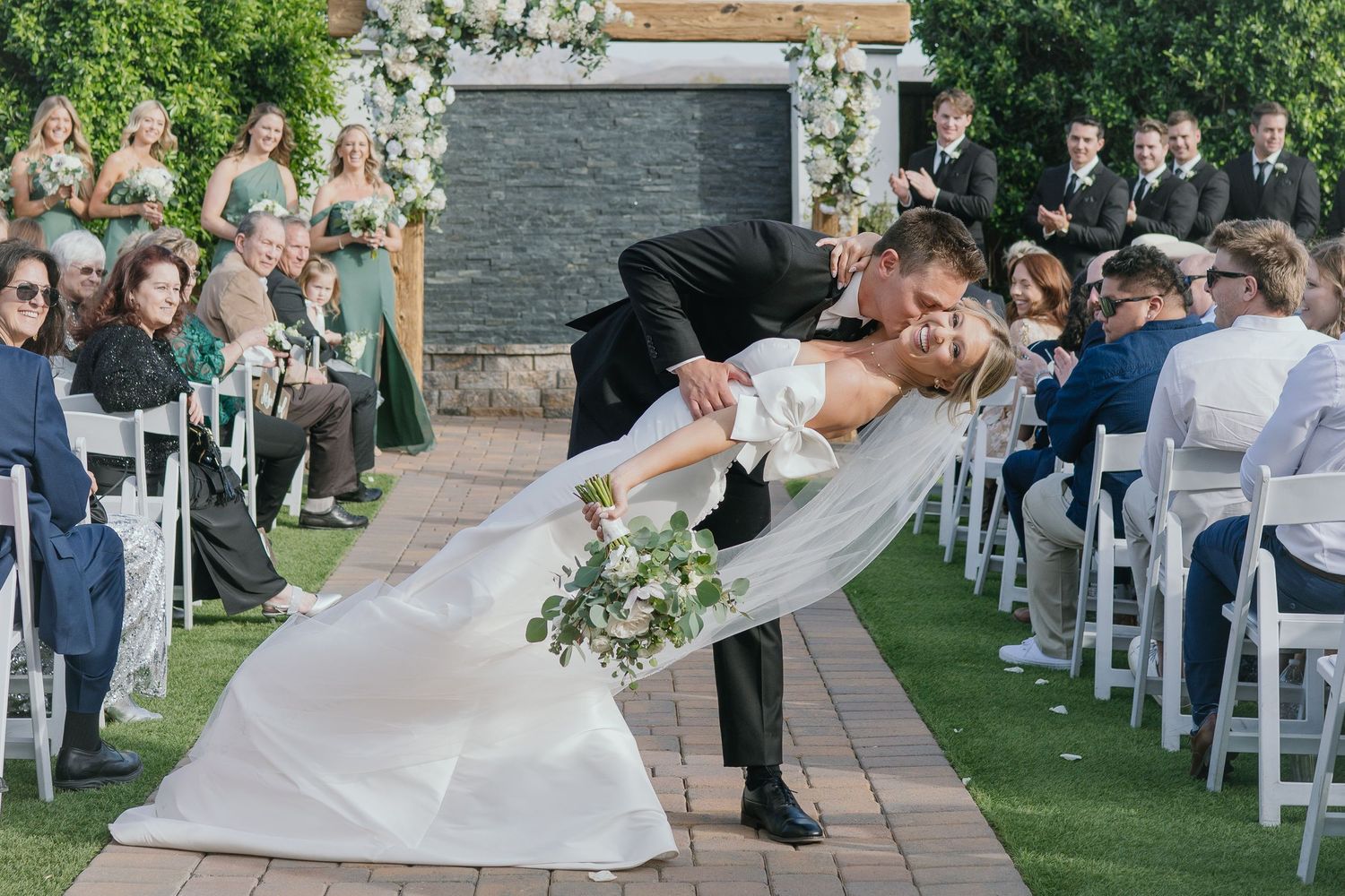Two people share a romantic dip kiss during an outdoor wedding ceremony while guests look on from white chairs.
