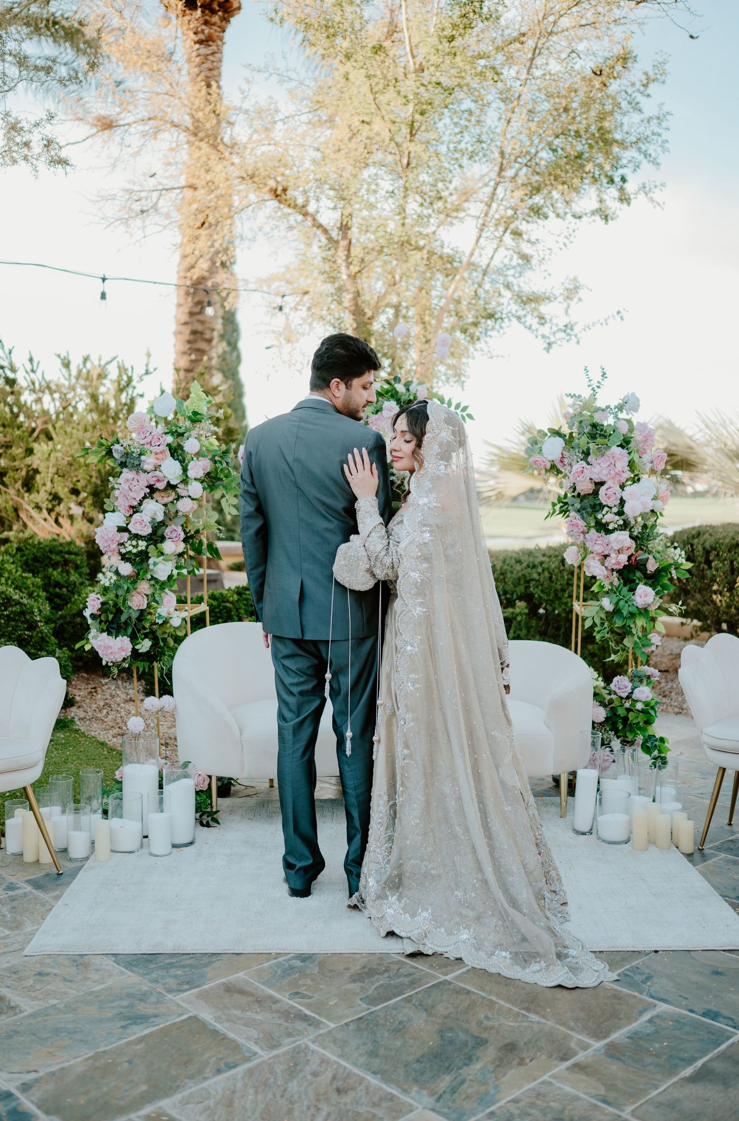 Romantic outdoor wedding ceremony with floral arches and candles decorating stone patio during golden hour.